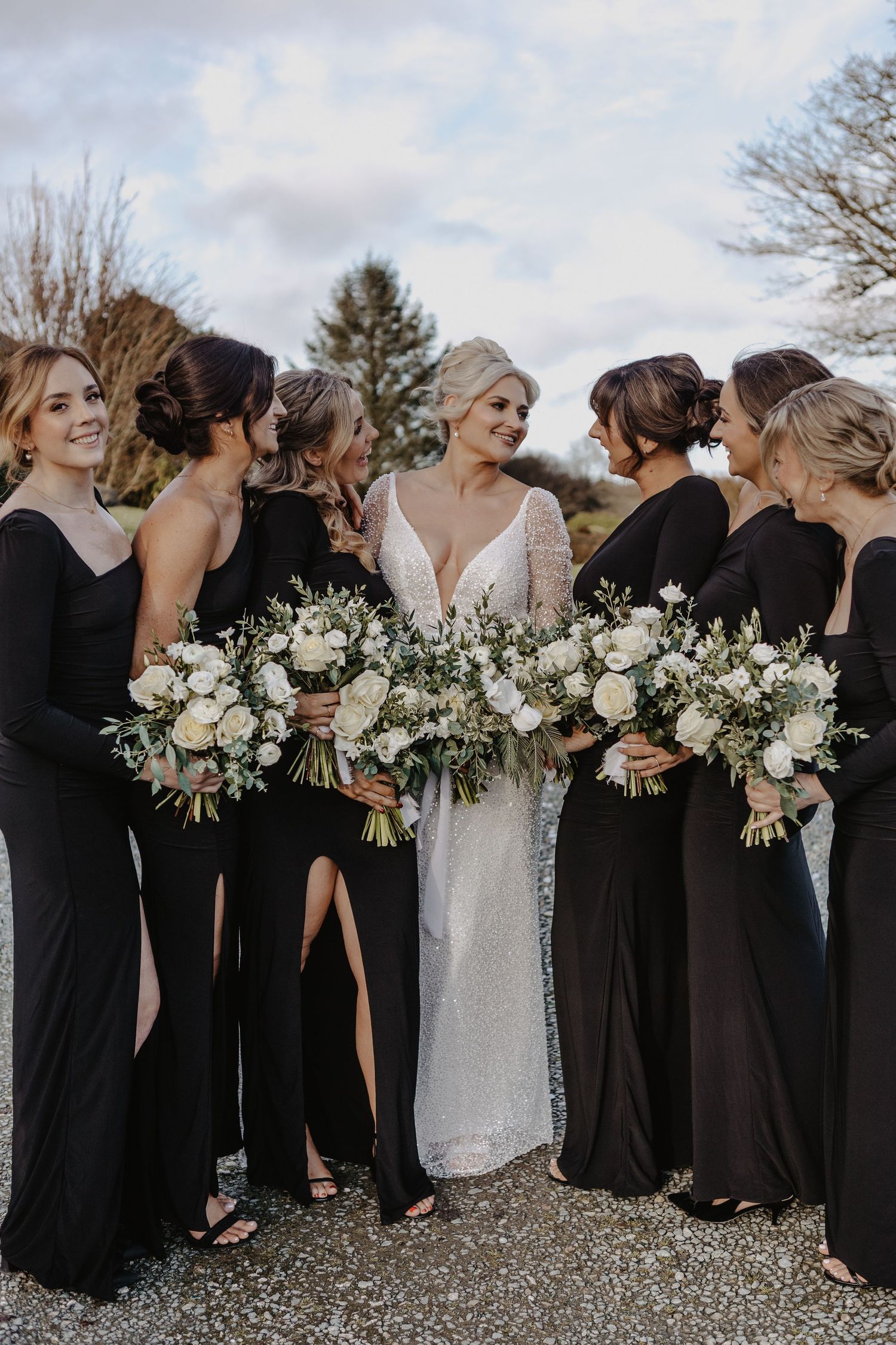 Bridal party in black dresses and white bouquets stands in line on gravel path during outdoor wedding.