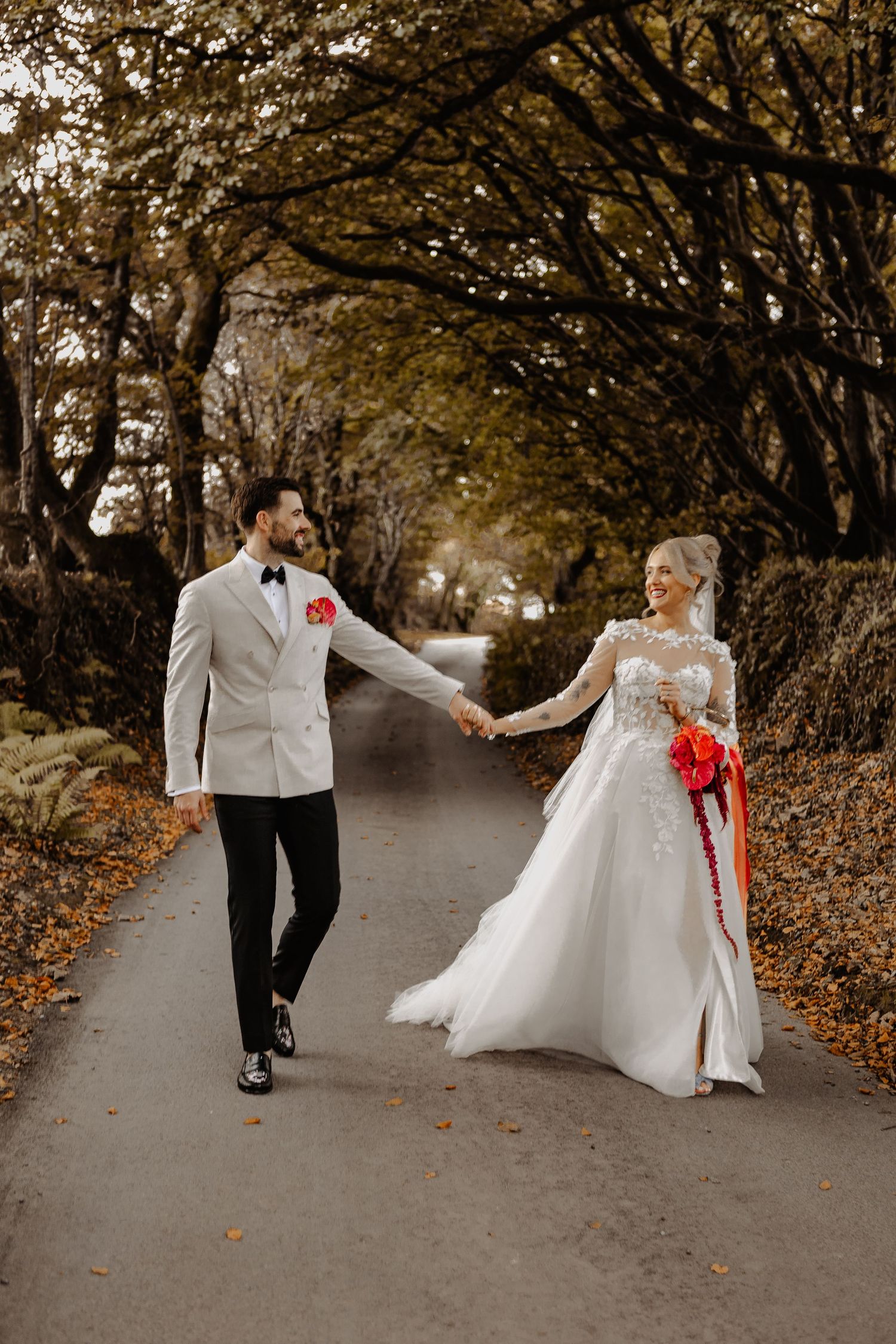 A couple in wedding attire walks together on a tree-lined autumn path with fallen leaves.