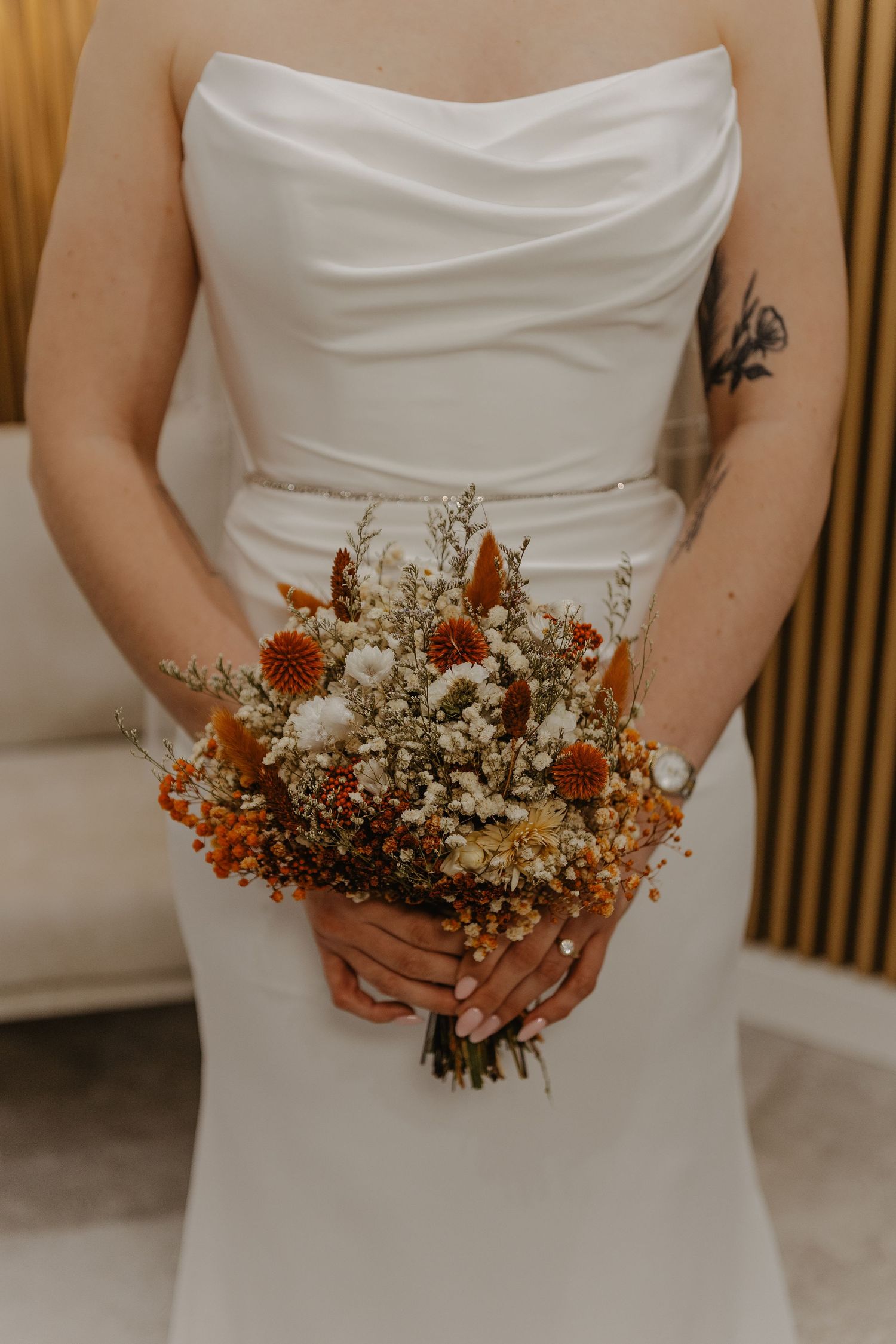 Close-up of bride holding rustic dried flower bouquet with orange and white blooms against white dress.