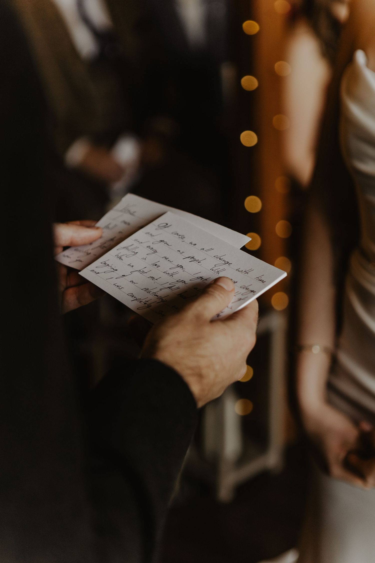 A hand holds a handwritten letter with elegant cursive writing against a dark background with soft bokeh lights.