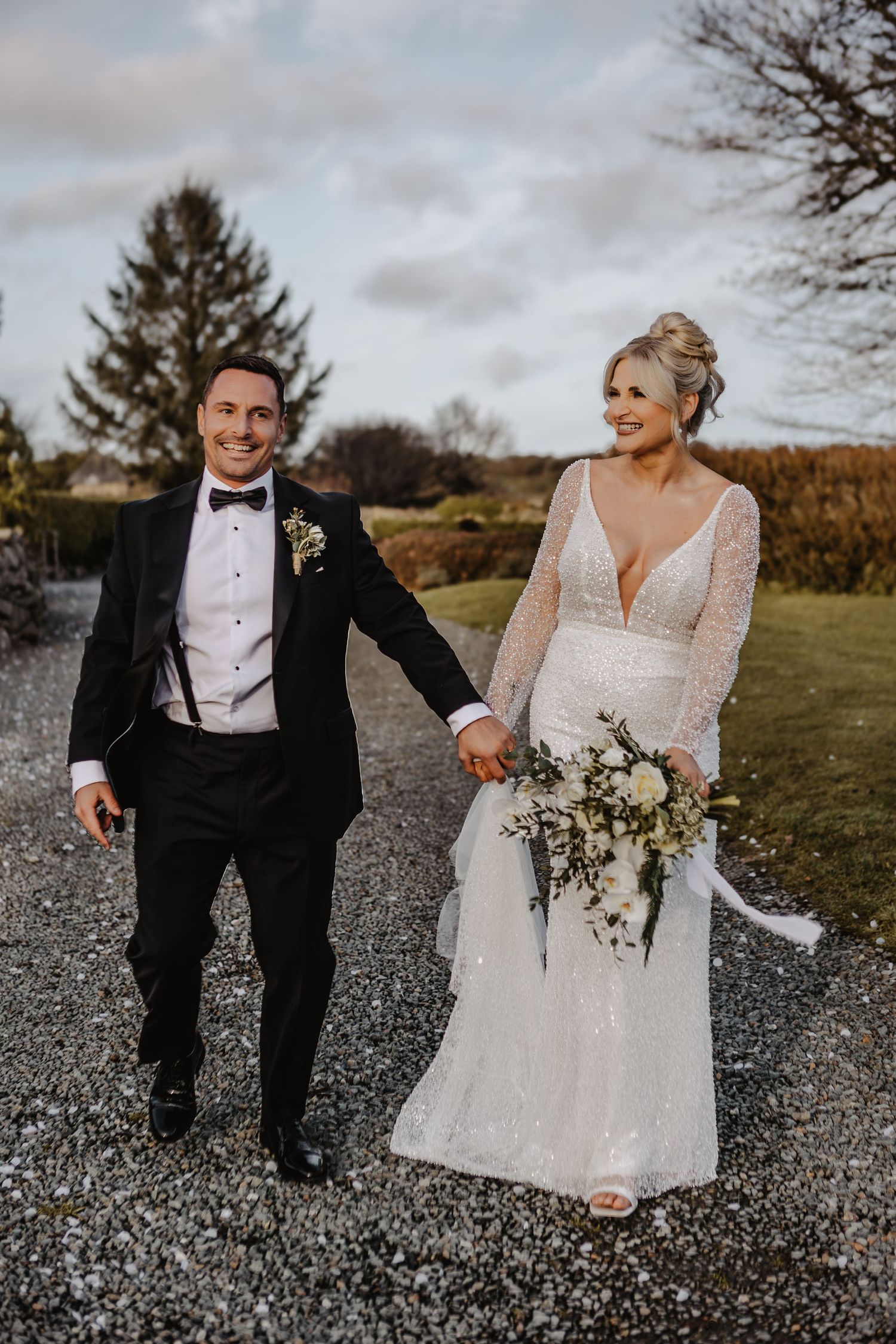 Wedding couple poses on gravel path in autumn landscape wearing traditional formal attire with white bouquet.