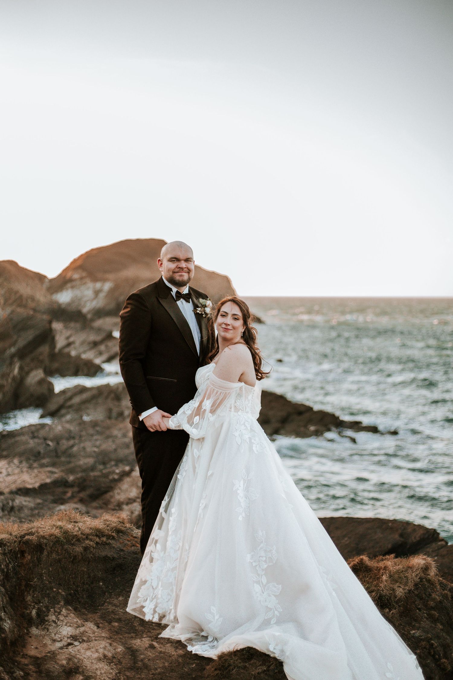A bride and groom pose on coastal rocks with ocean waves and cliffs in the background on their wedding day.