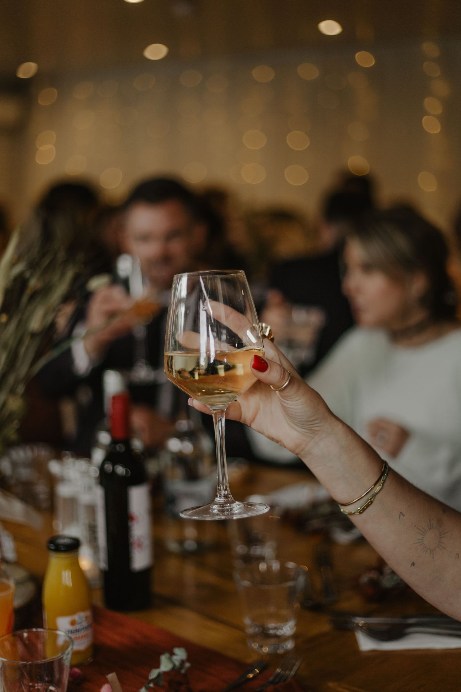Hand holding a glass of white wine at a celebratory dinner table with bokeh lights in background.
