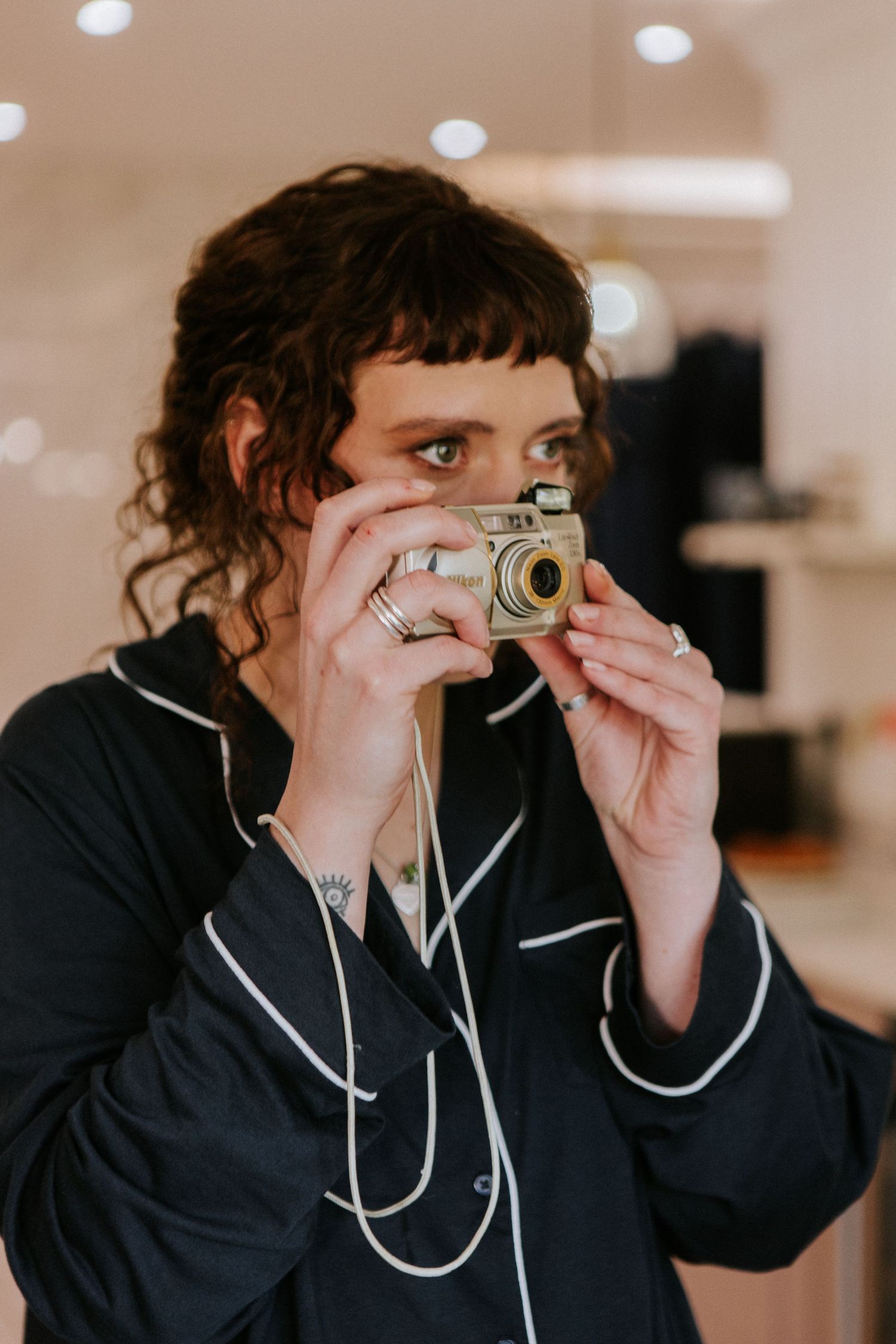 Young woman with curly hair photographing with a vintage gold camera in a softly lit indoor setting.