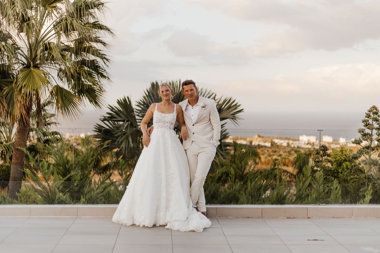 Bride in a white gown and groom in a cream suit embrace on a terrace with palm trees and a scenic ocean view at sunset.