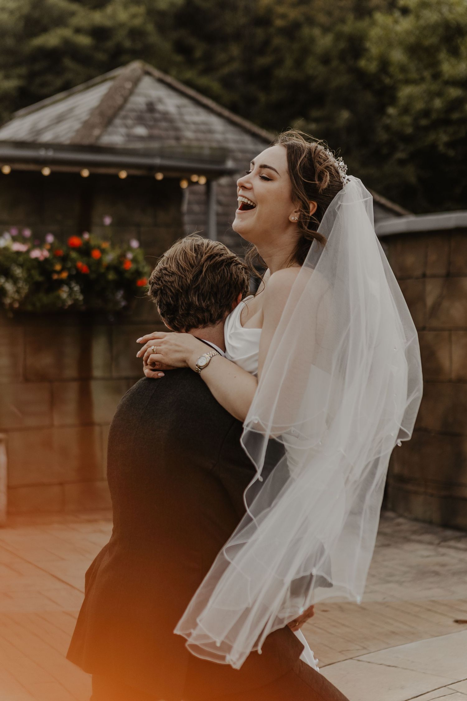 A bride in a flowing veil laughs joyfully during an outdoor wedding photoshoot at sunset.