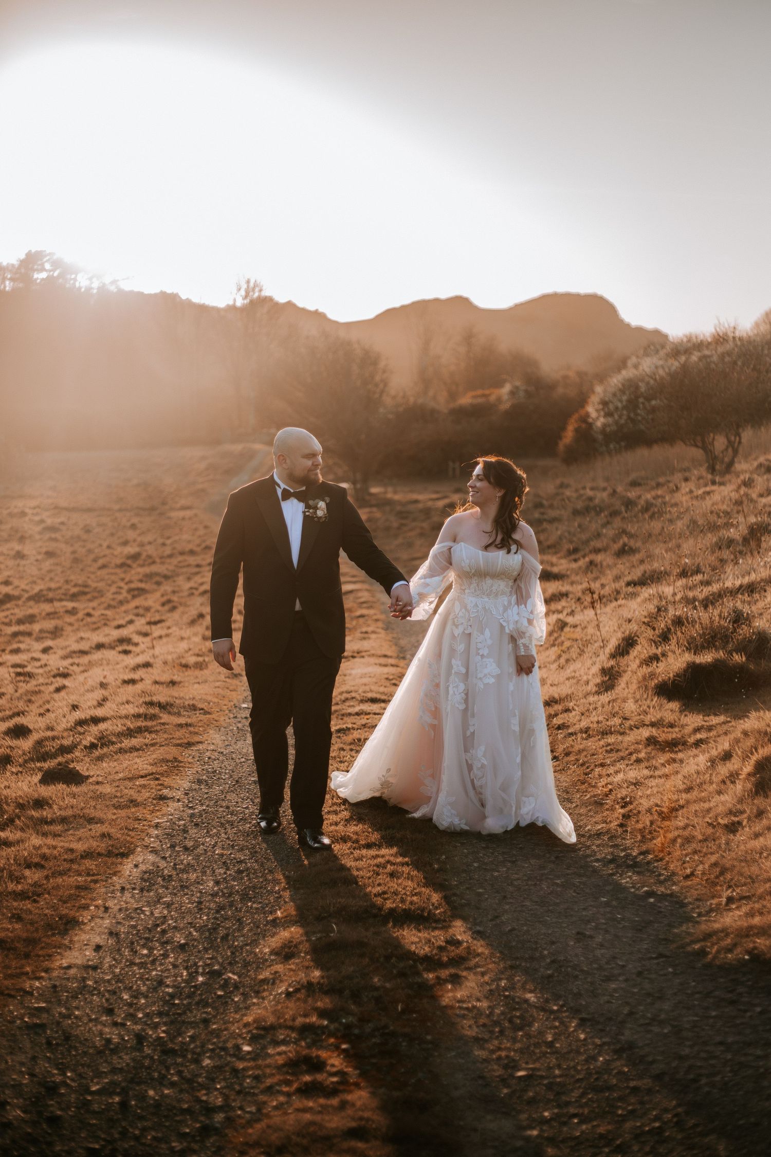 Bride and groom holding hands walking on a path at golden hour with scenic hills in the background.