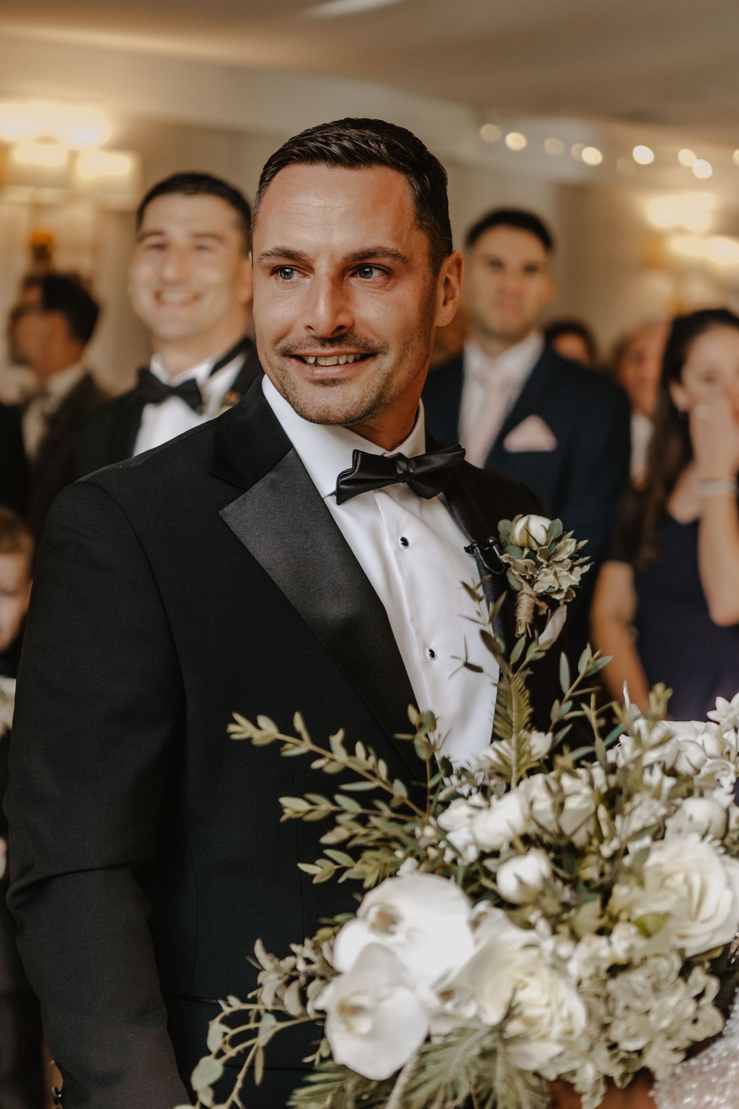 Groomsmen in black tuxedos and bow ties stand together with white floral boutonnieres at an elegant wedding.