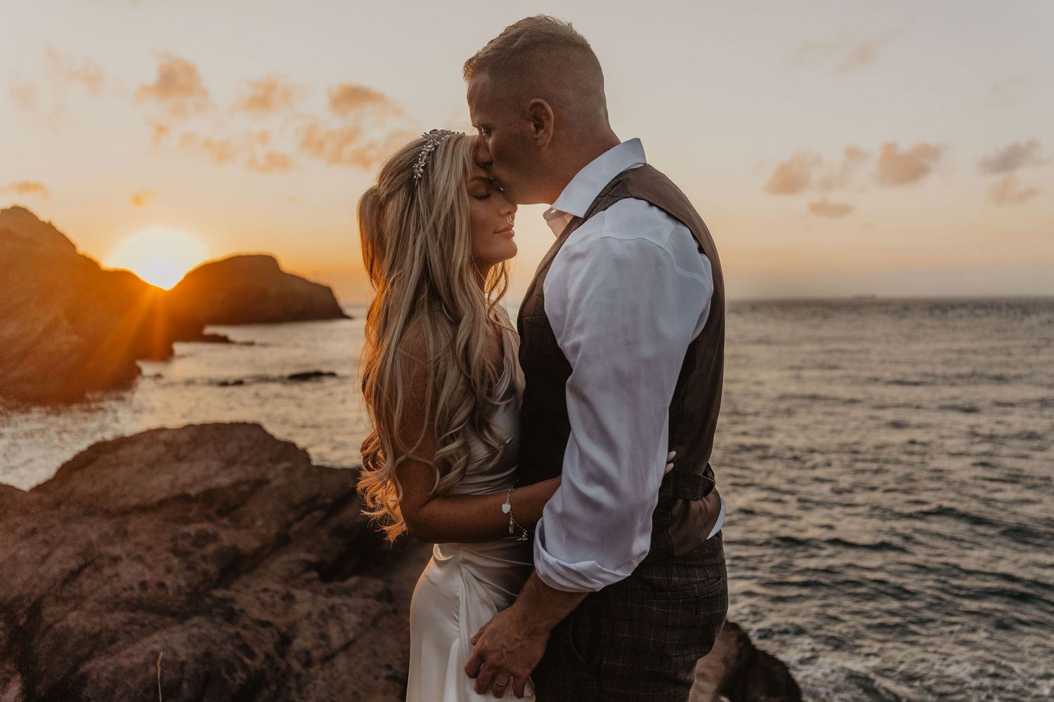 Romantic couple embracing on beach during golden sunset with ocean waves and cliffs in background.