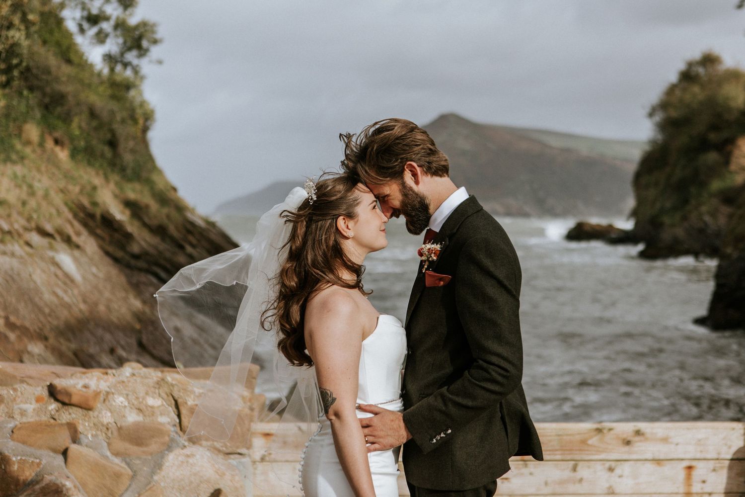 A couple shares an intimate moment on a rocky coastline with dramatic cliffs and stormy waters in the background.