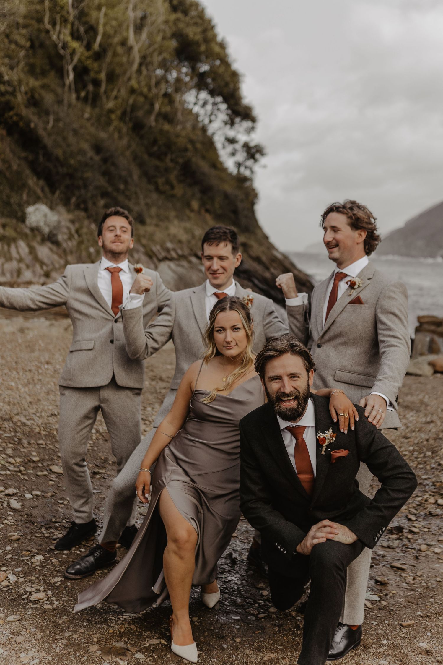 Wedding party in gray suits and rust dresses pose casually on rocky beach.