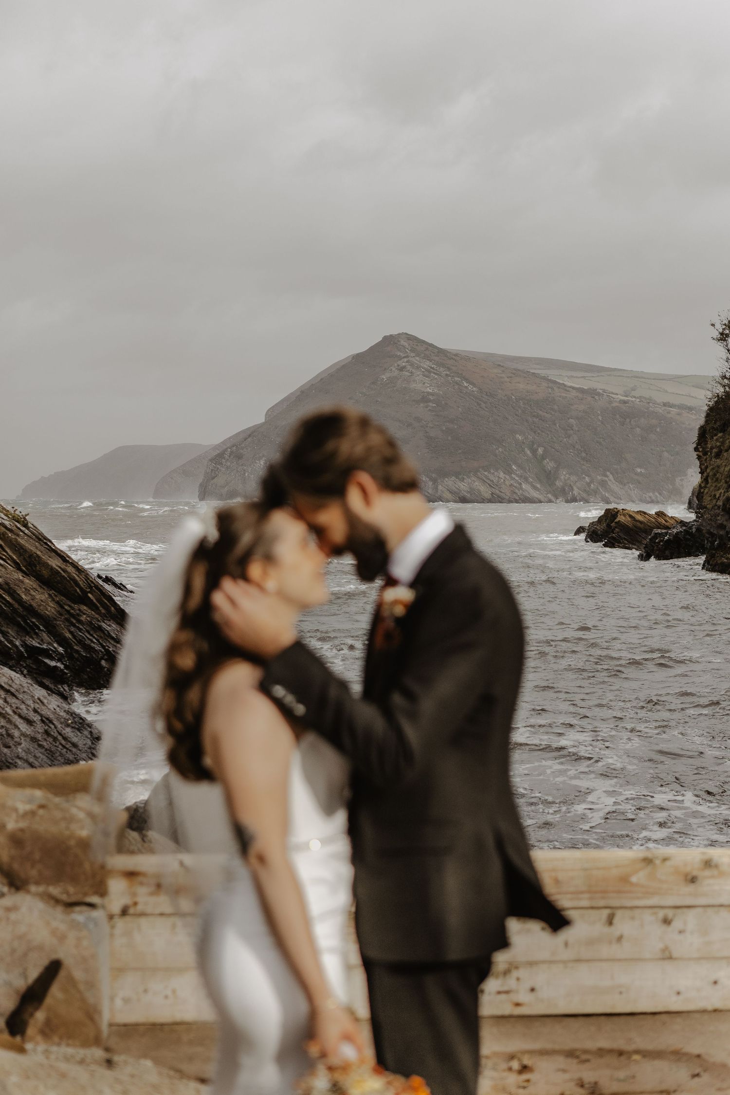 A couple shares a romantic moment on a rocky coastline with dramatic cliffs and crashing waves in the background.
