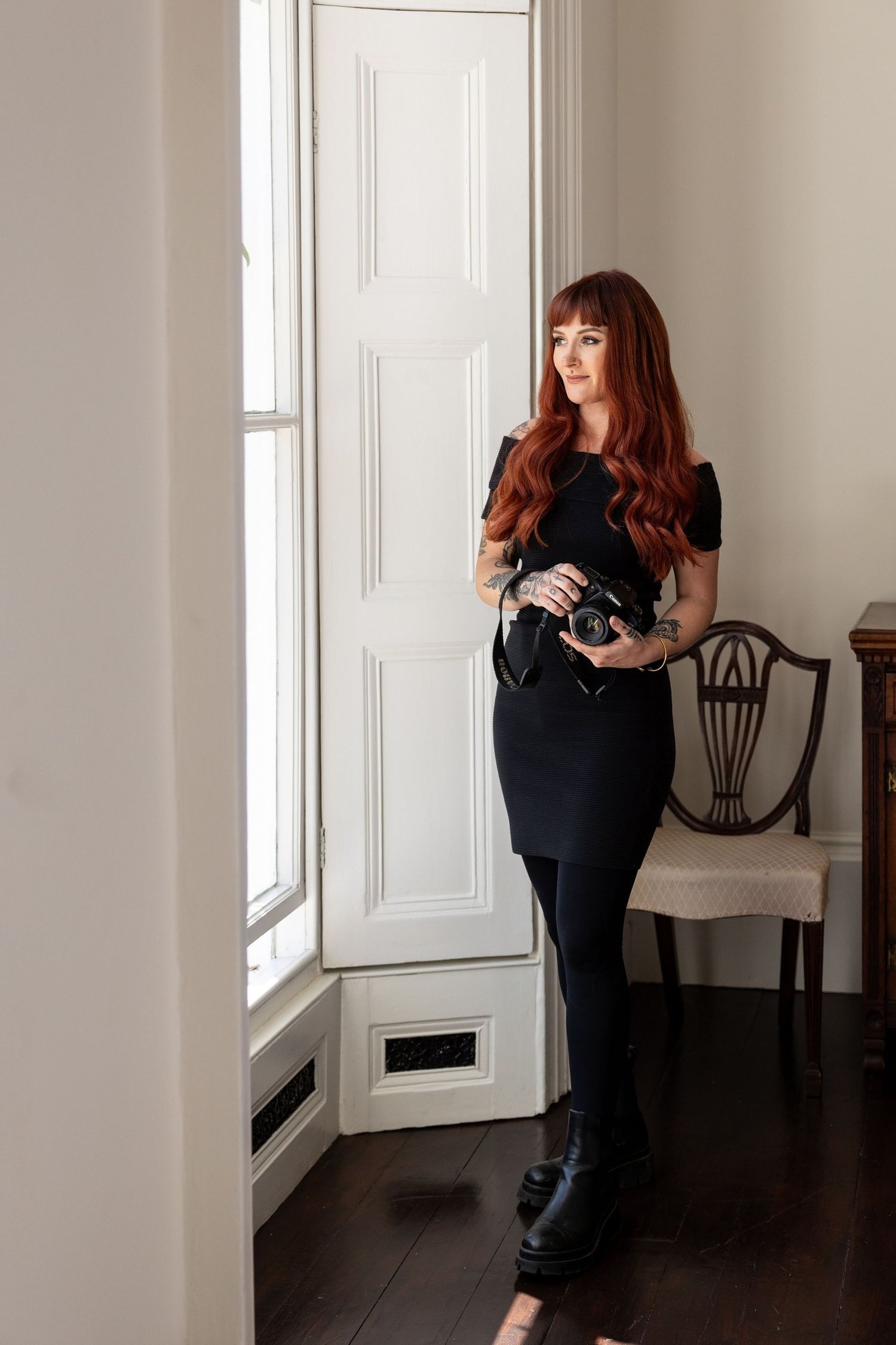 Professional photographer in black dress holding camera in bright interior studio setting.
