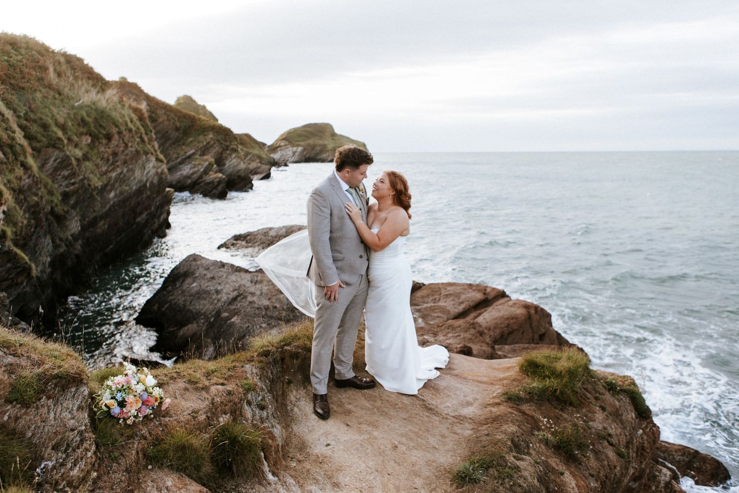 A couple in wedding attire shares a romantic moment on a dramatic coastal cliff overlooking the ocean.