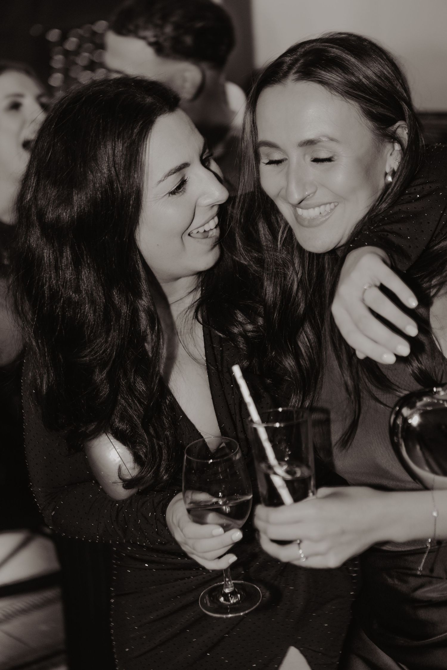 Two friends share a candid moment laughing together while holding drinks at a social gathering in black and white.