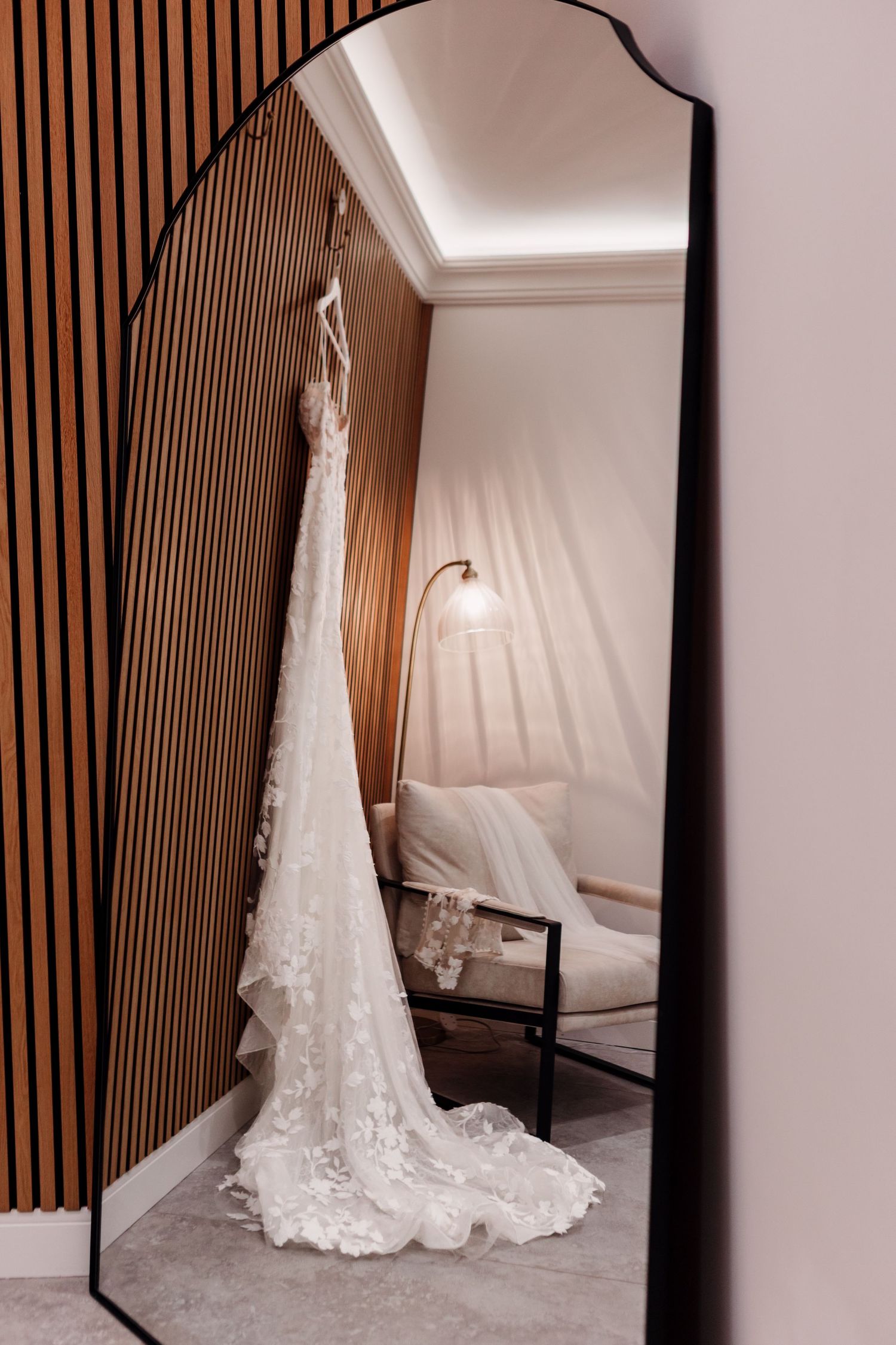 A white lace wedding dress hangs elegantly beside a full-length mirror in a dressing room with wood-paneled walls.