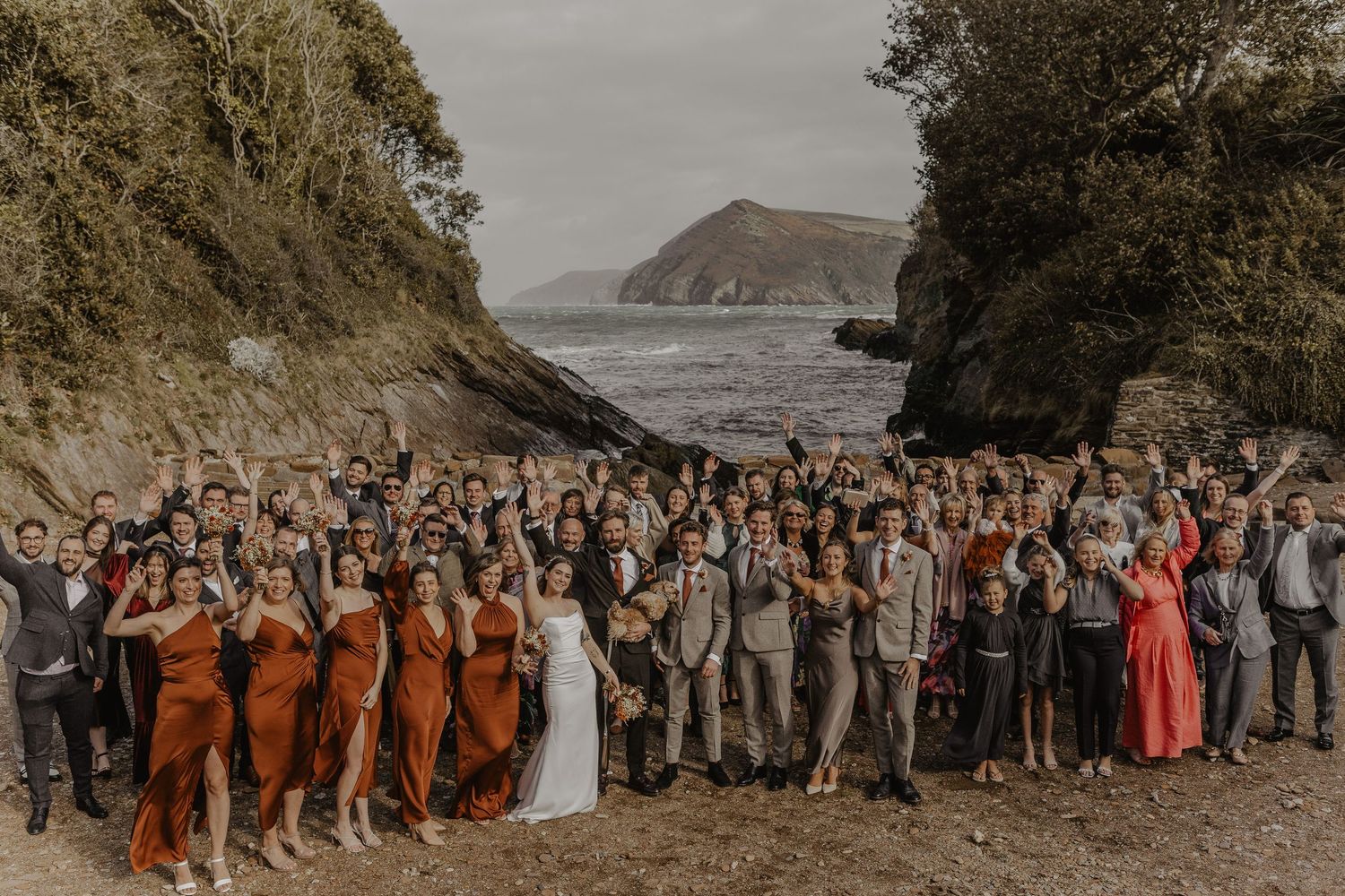 Large wedding group photo with rust-colored bridesmaid dresses against a coastal backdrop.