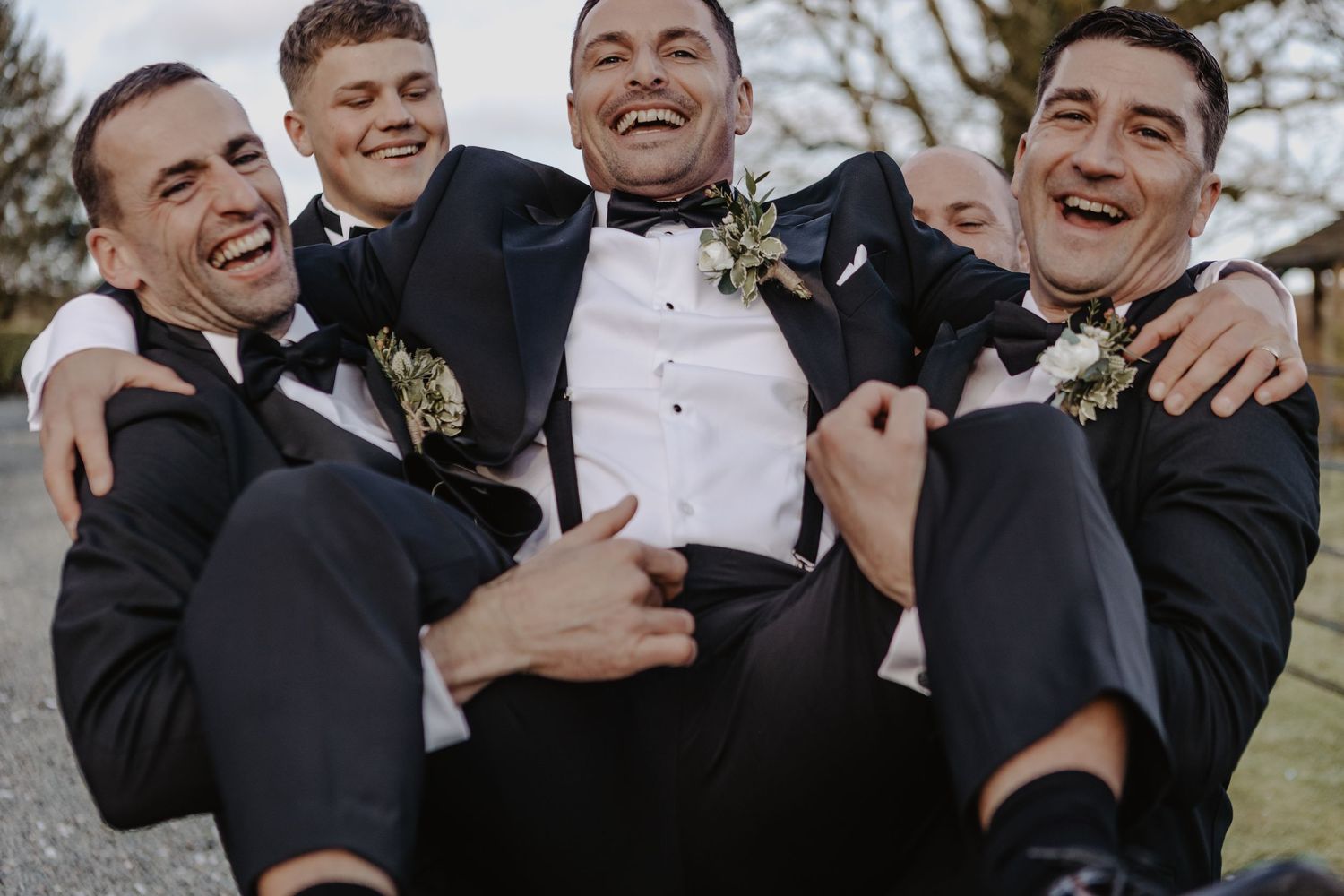 Groomsmen in black tuxedos share laughs and embrace during outdoor wedding photo session.
