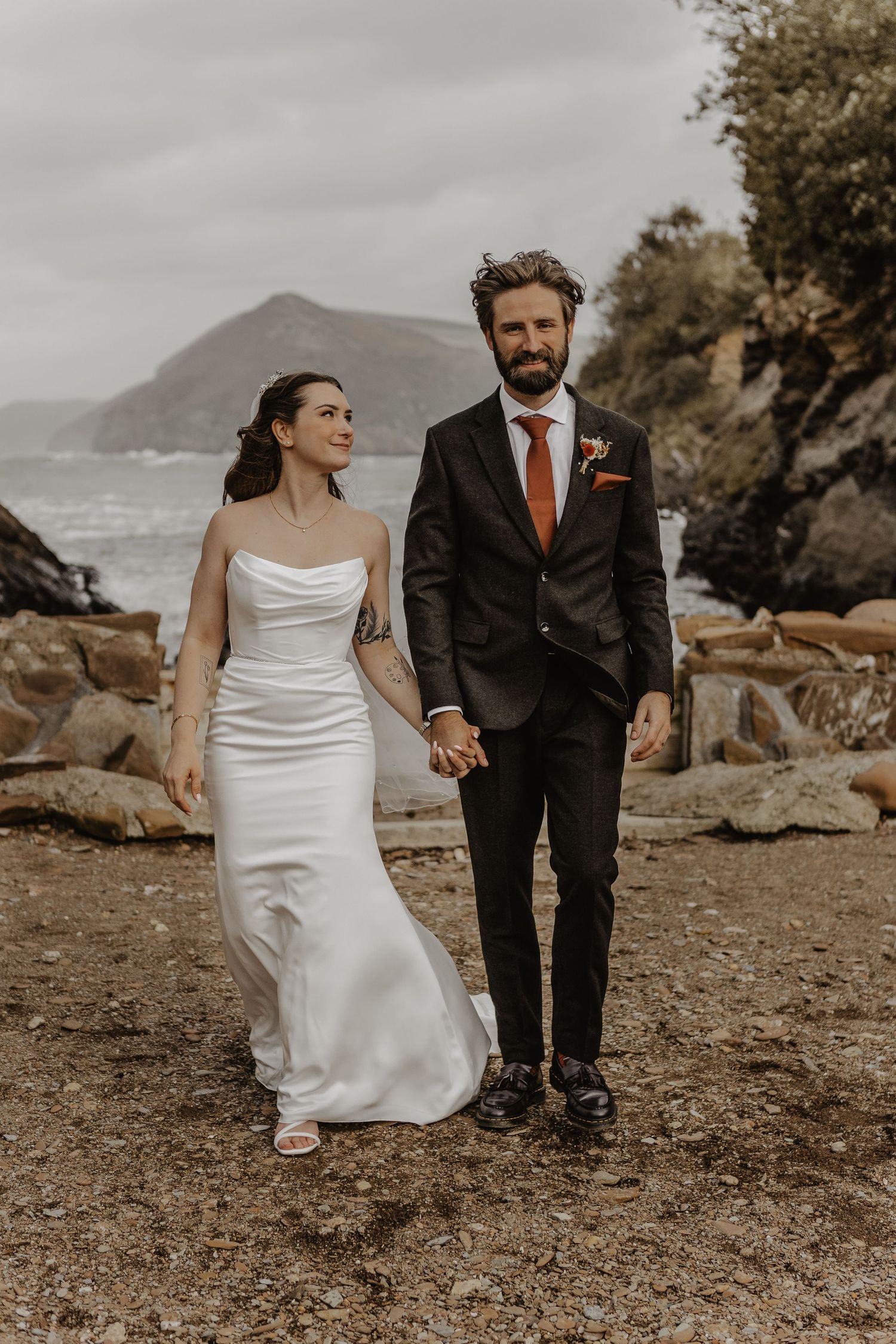 Couple in wedding attire walking along a rocky coastal landscape with mountains in the background.
