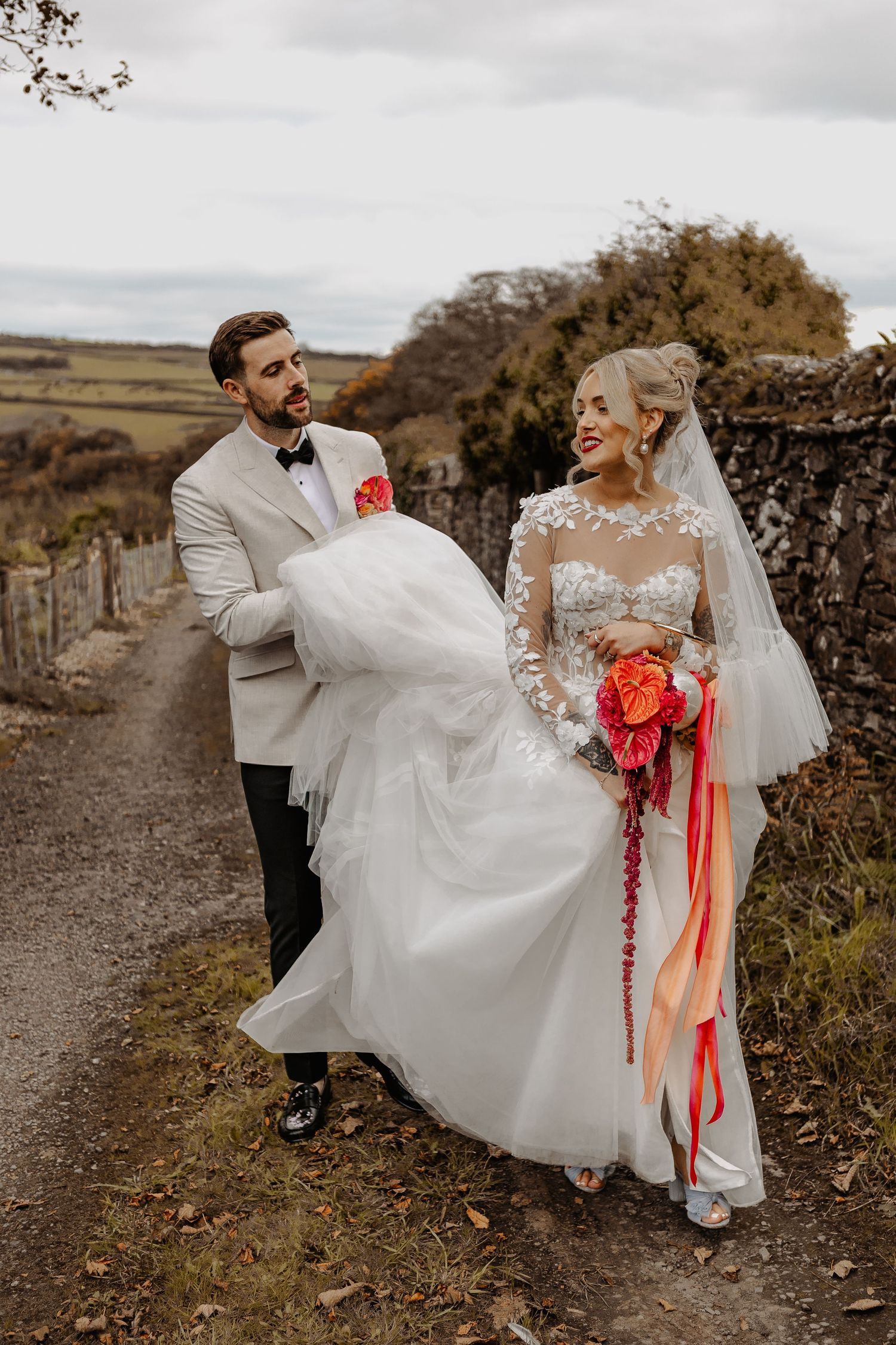 Joyful couple in wedding attire dances and twirls along a rustic stone path surrounded by autumn foliage.