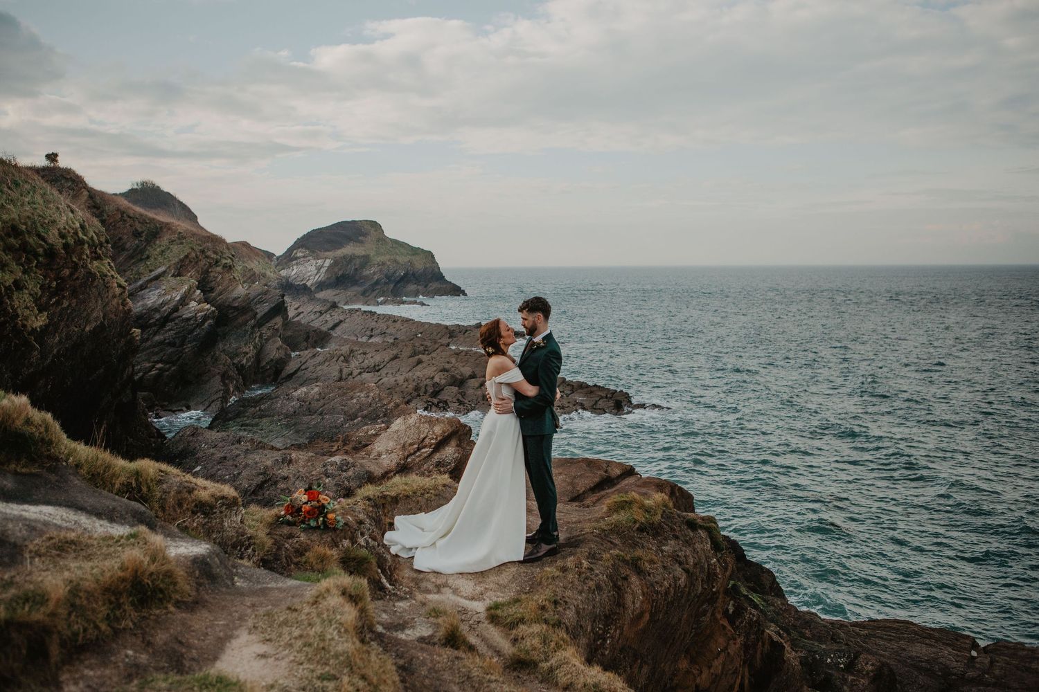 A couple stands on dramatic coastal cliffs overlooking the ocean during a scenic outdoor wedding ceremony.