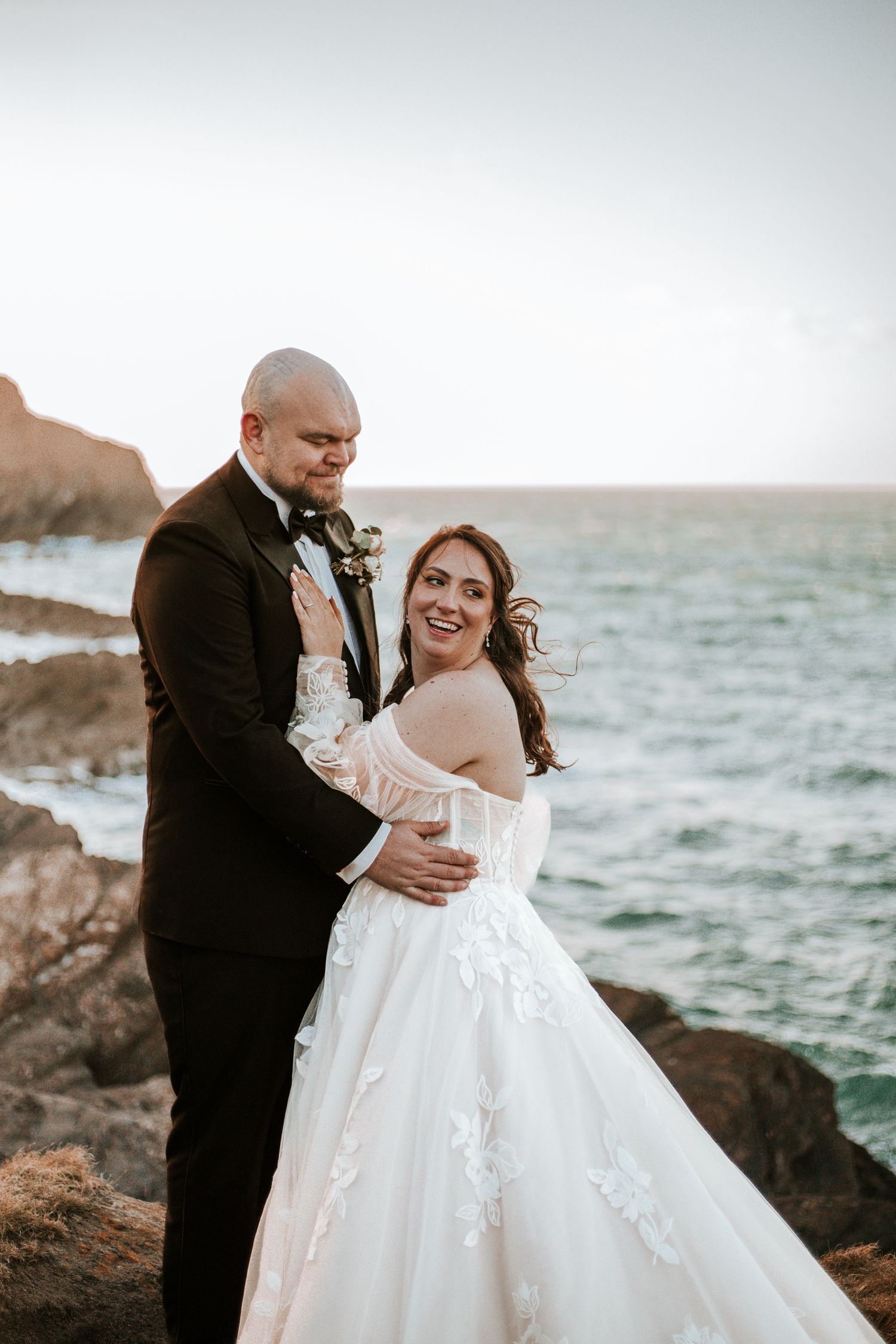 Bride in off-shoulder lace gown and groom in black tuxedo embrace on rocky coastal cliffs at sunset.