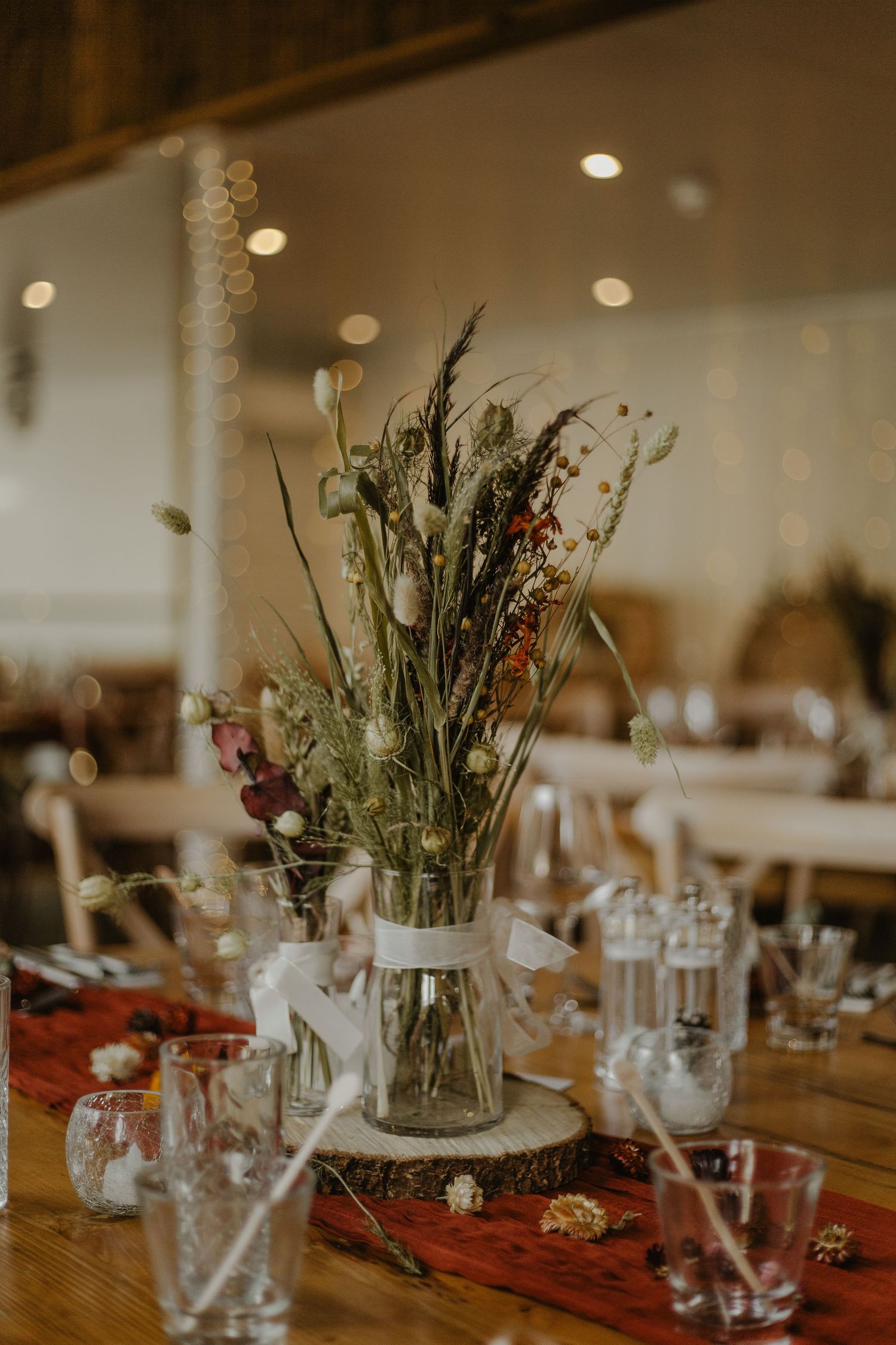 Rustic wedding reception table settings with wildflower centerpieces in glass vases on wooden tables with red runners.