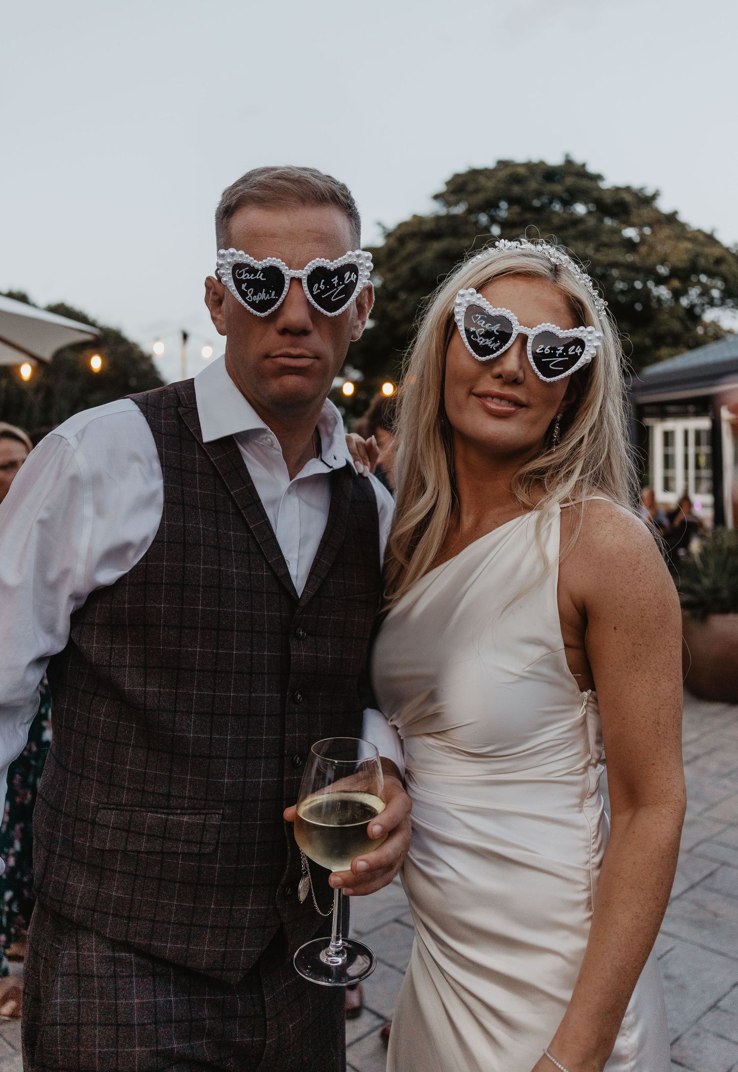 A couple wearing sunglasses and formal attire pose together at an outdoor wedding reception with string lights visible.