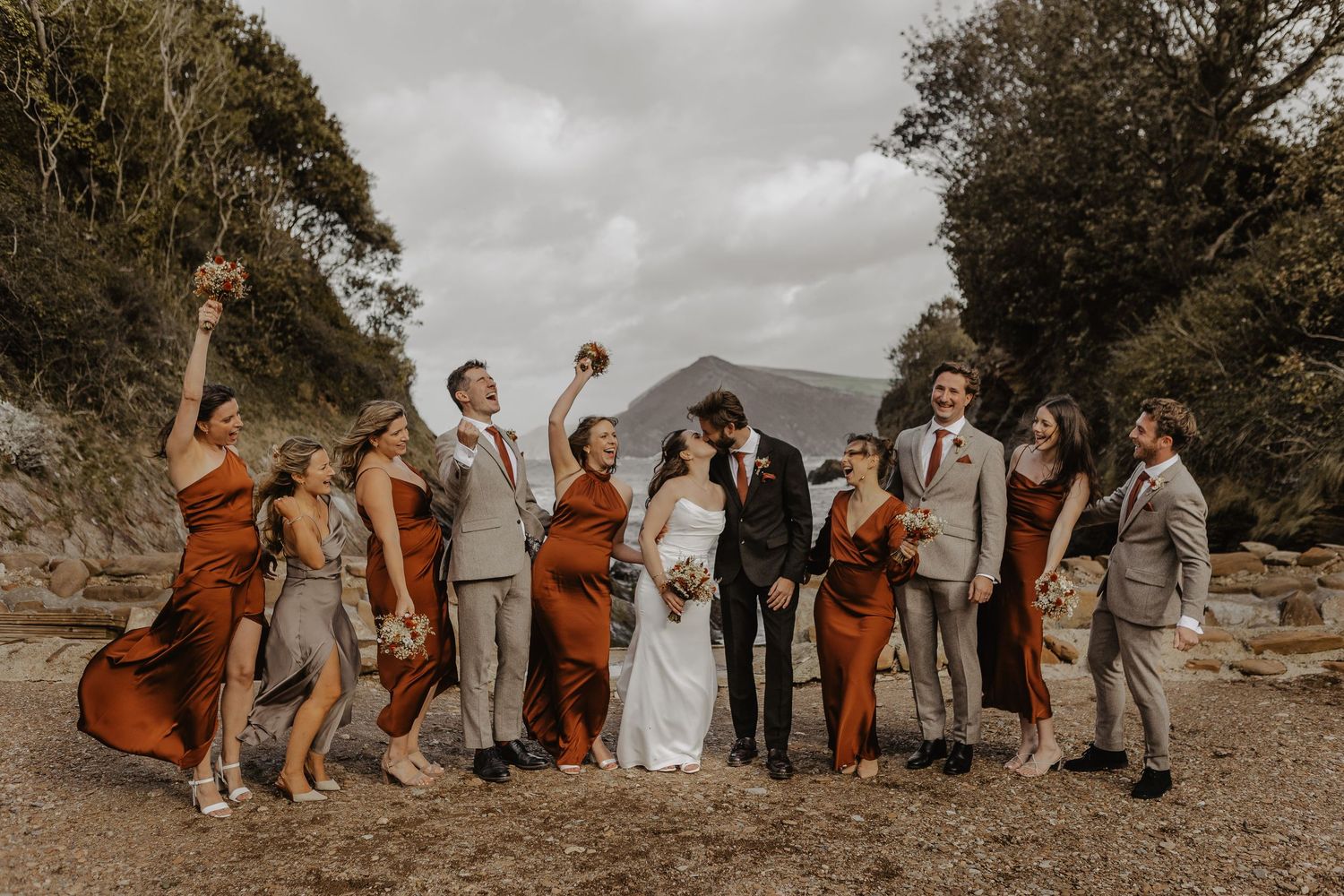 Wedding party in gray and rust orange attire poses together on a scenic outdoor cliff overlooking mountains.