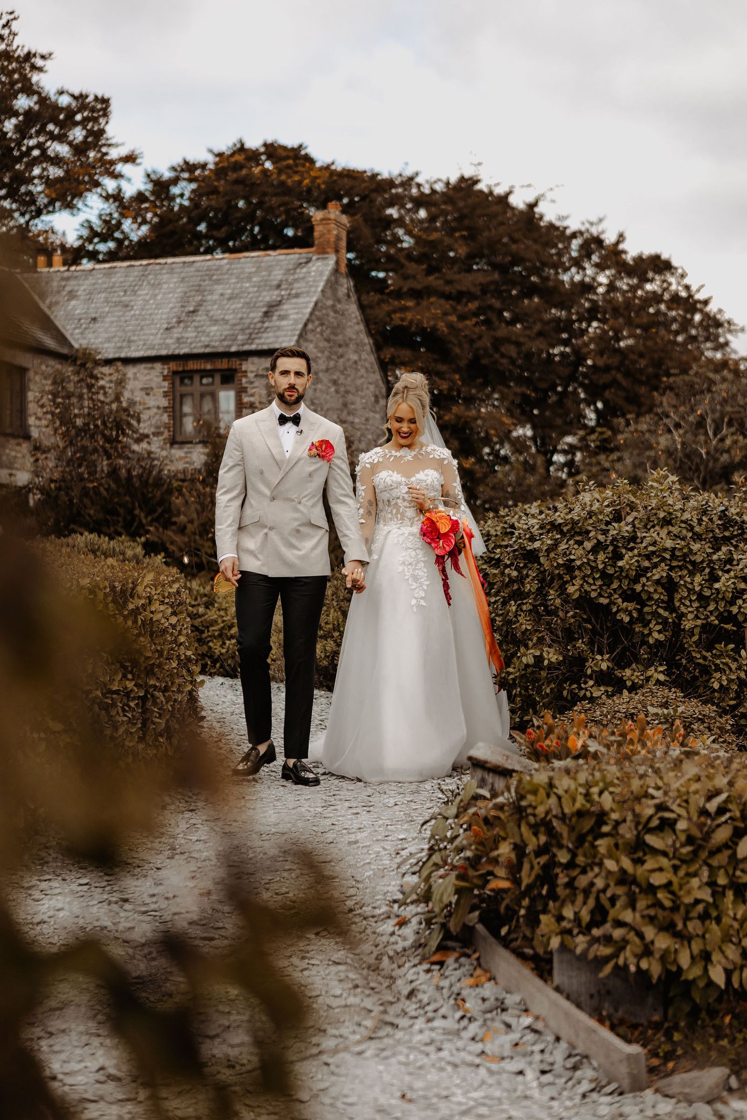 A romantic wedding couple walks along a stone path surrounded by autumn foliage on an overcast day.