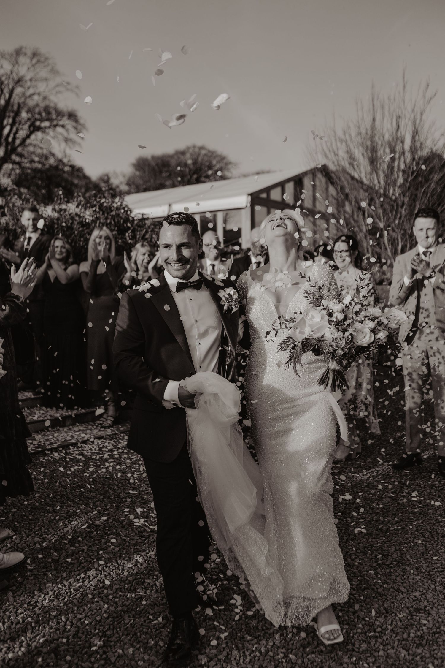 A black and white wedding photo showing a couple joyfully walking through confetti surrounded by celebrating guests.