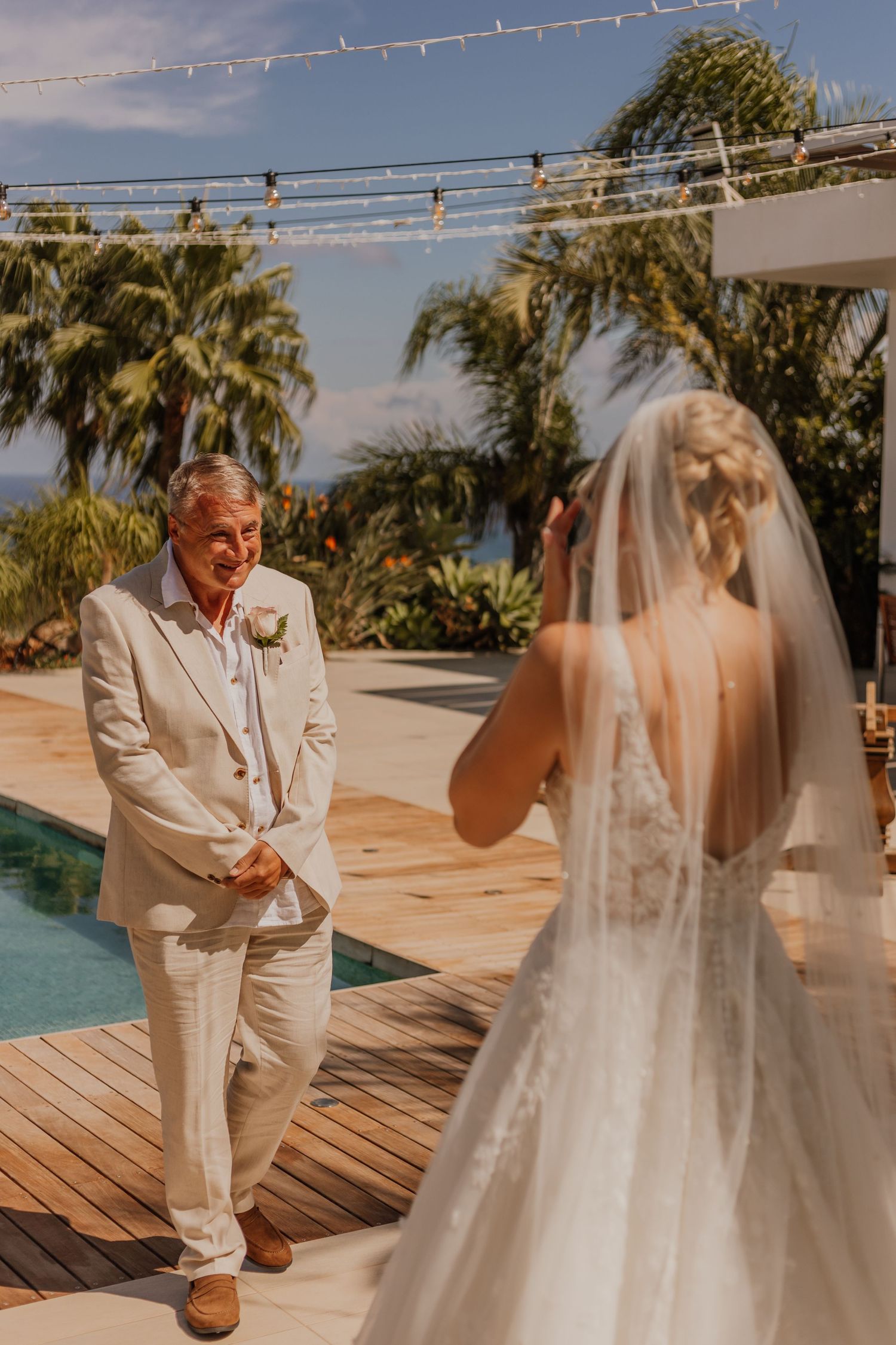 Bride in a lace gown and long veil photographs a smiling groom in a cream suit beside a poolside deck with palm trees.