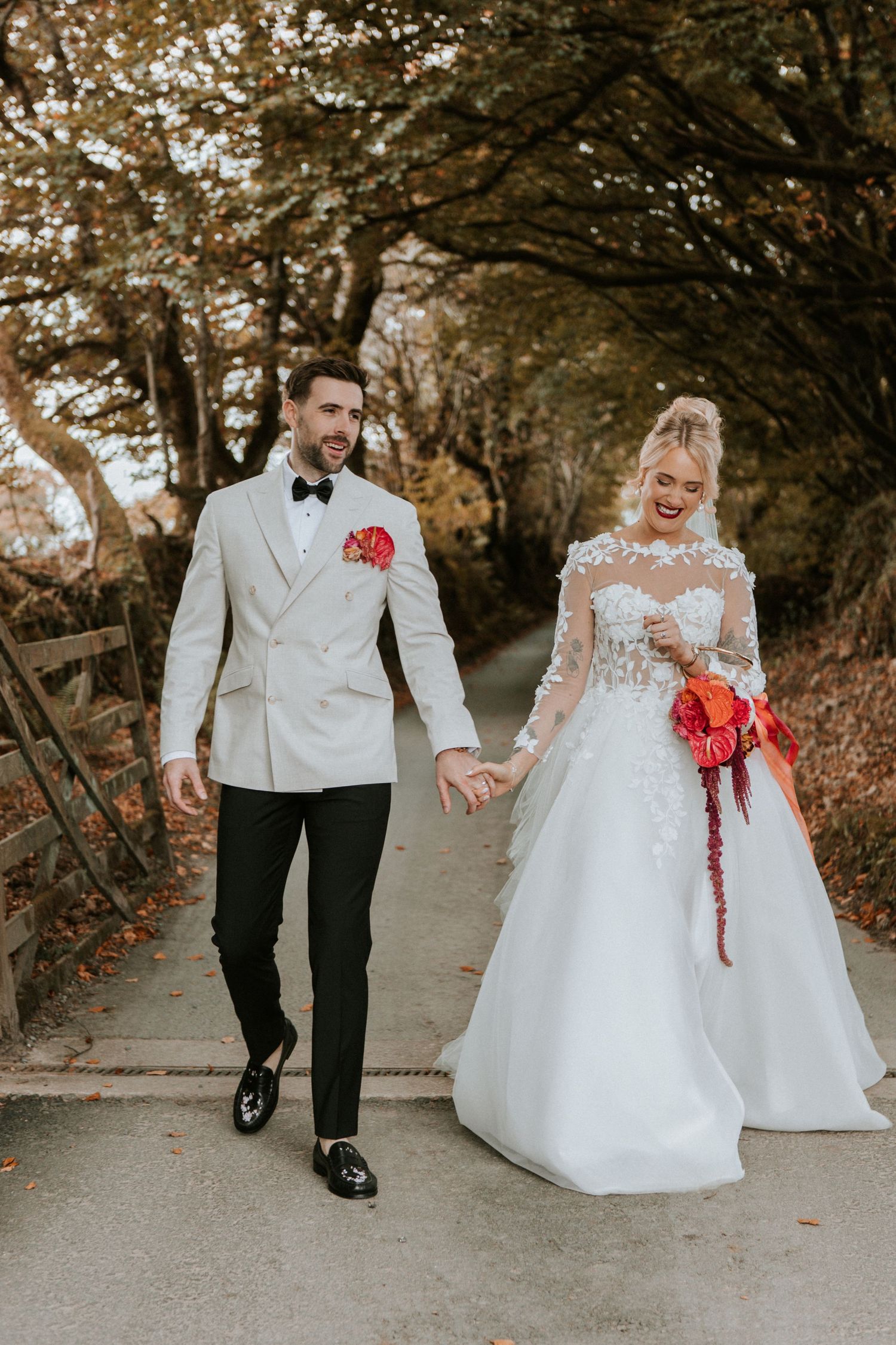 A couple walking together on a tree-lined path, with the bride in a white gown and groom in formal attire.