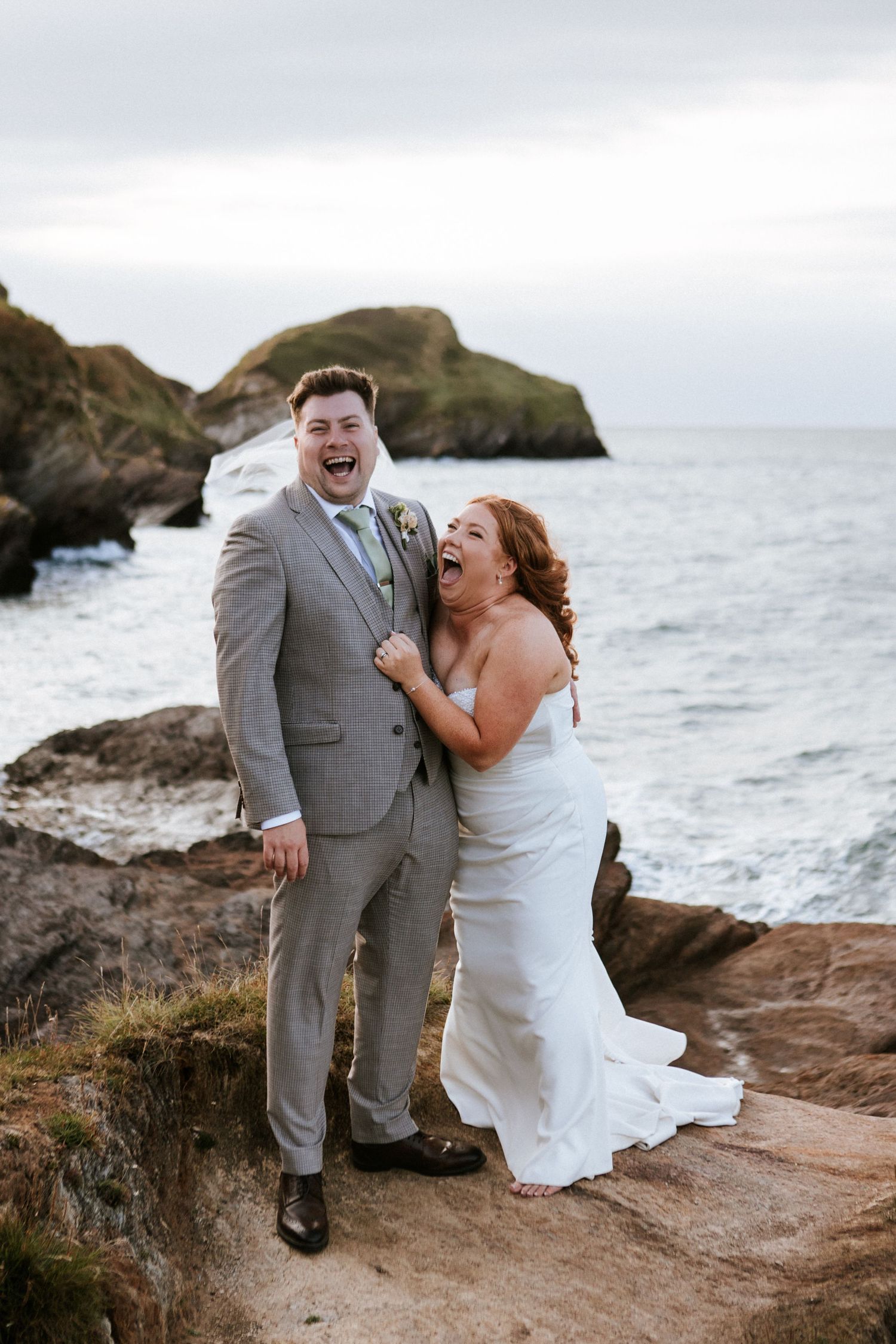 A couple shares joyful candid moments on a rocky coastal cliff overlooking the ocean during their wedding celebration.