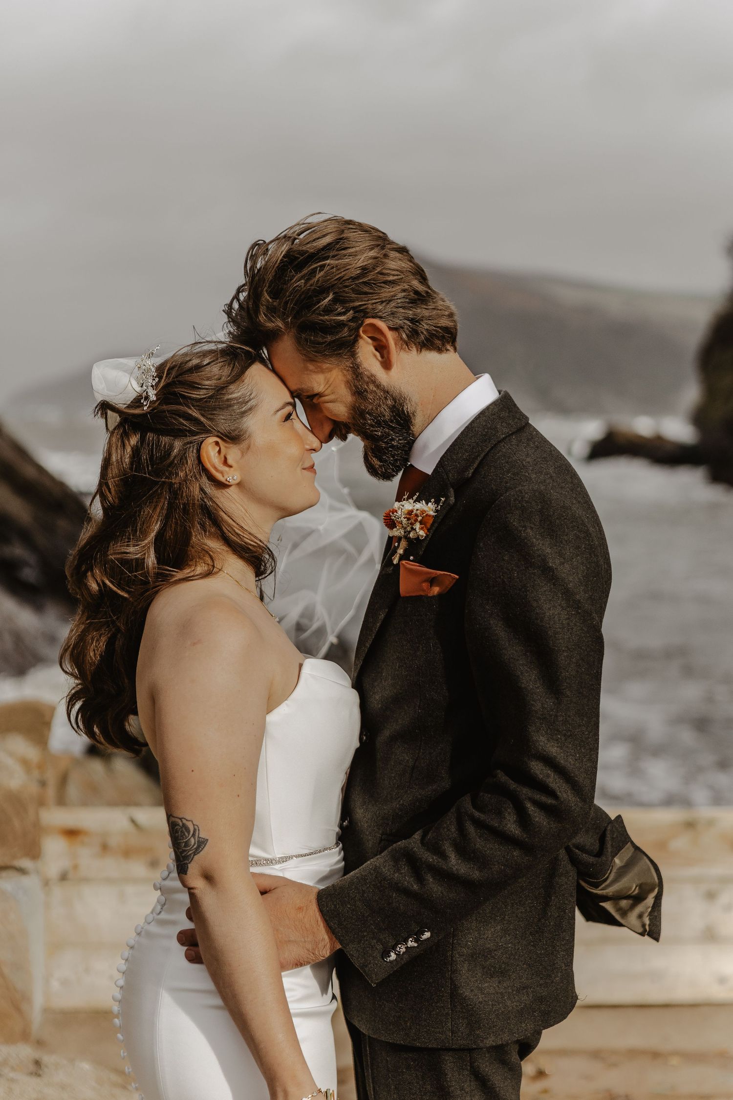 Intimate wedding portrait of couple embracing on rocky coastline with moody grey skies.