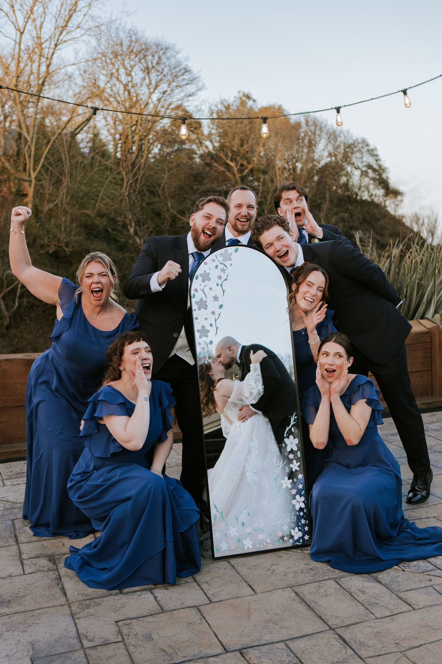 Wedding party in navy blue poses excitedly around a mirror reflecting the bride and groom kissing outdoors.