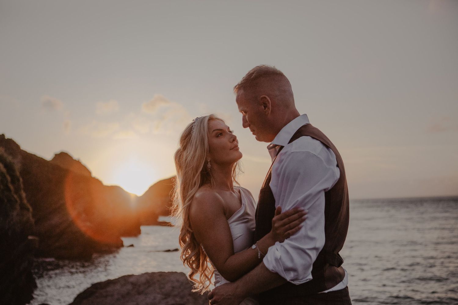 A romantic couple embraces on a beach at sunset with ocean waves and golden light in the background.