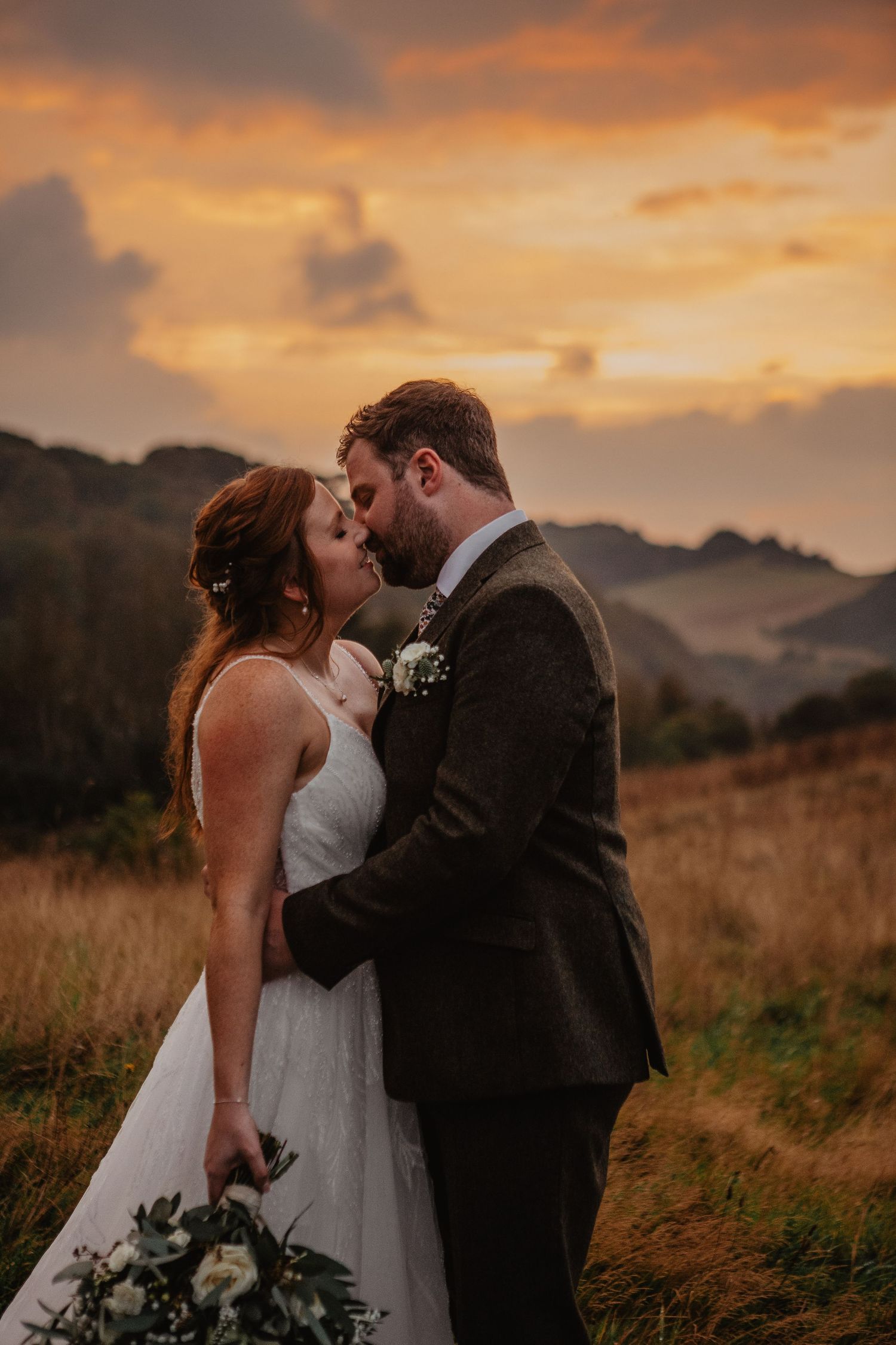 A couple shares a romantic kiss at sunset in a mountainous field with dramatic orange and purple skies.