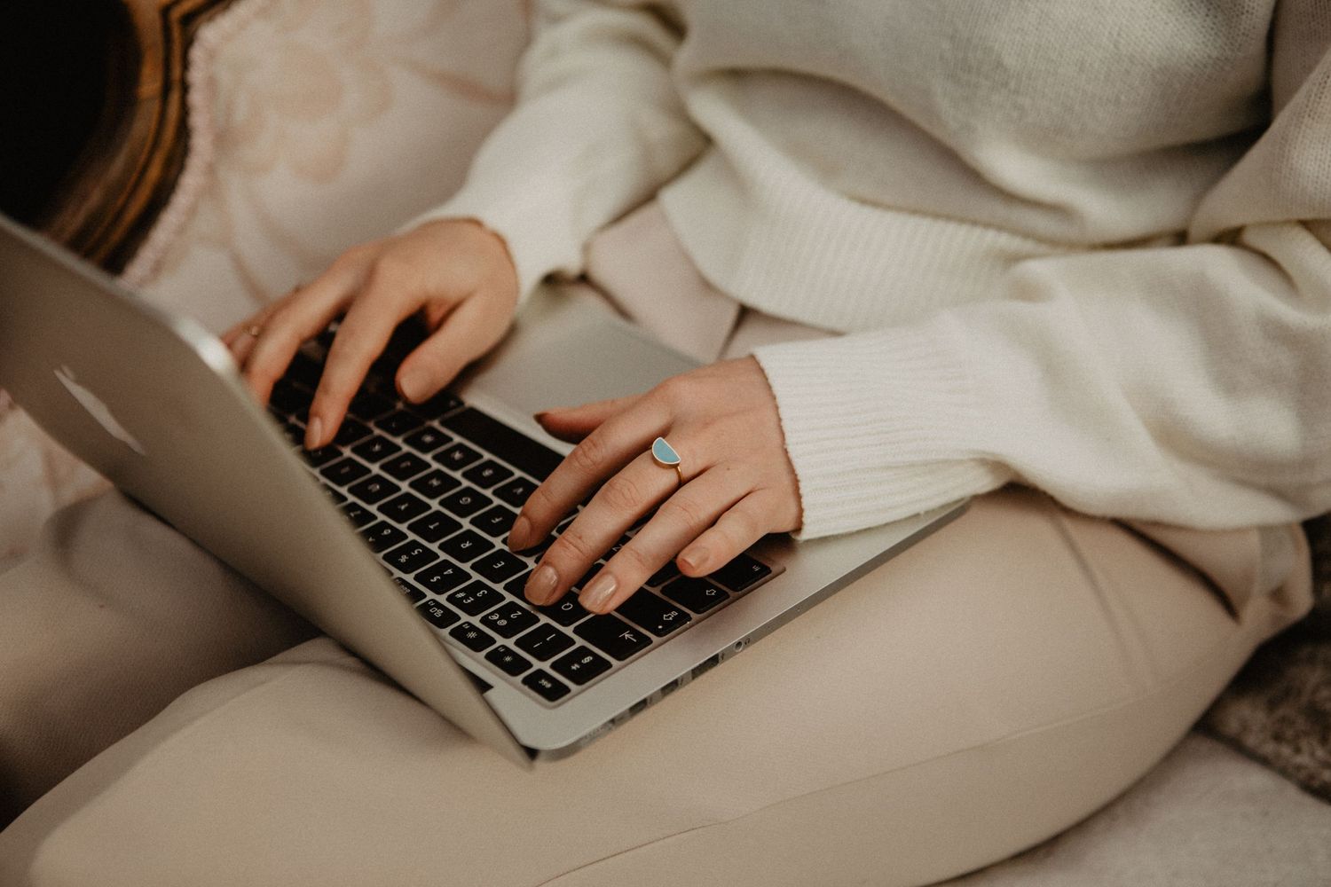 Person in cream sweater typing on MacBook laptop while sitting comfortably on couch.