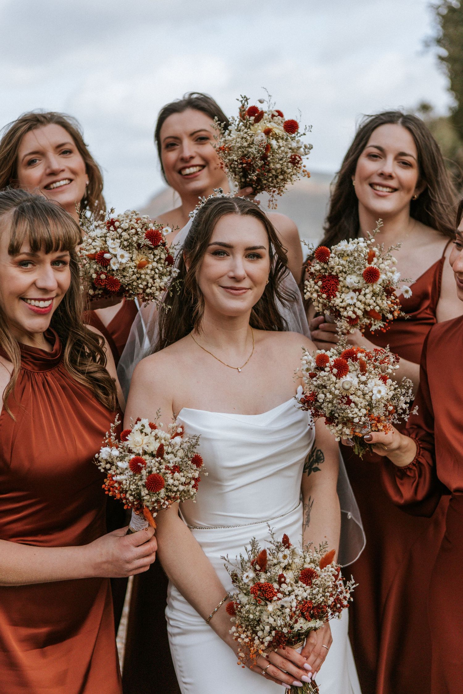 A bridal party in rust-colored dresses holds beautiful bouquets of red and white flowers against an overcast sky.