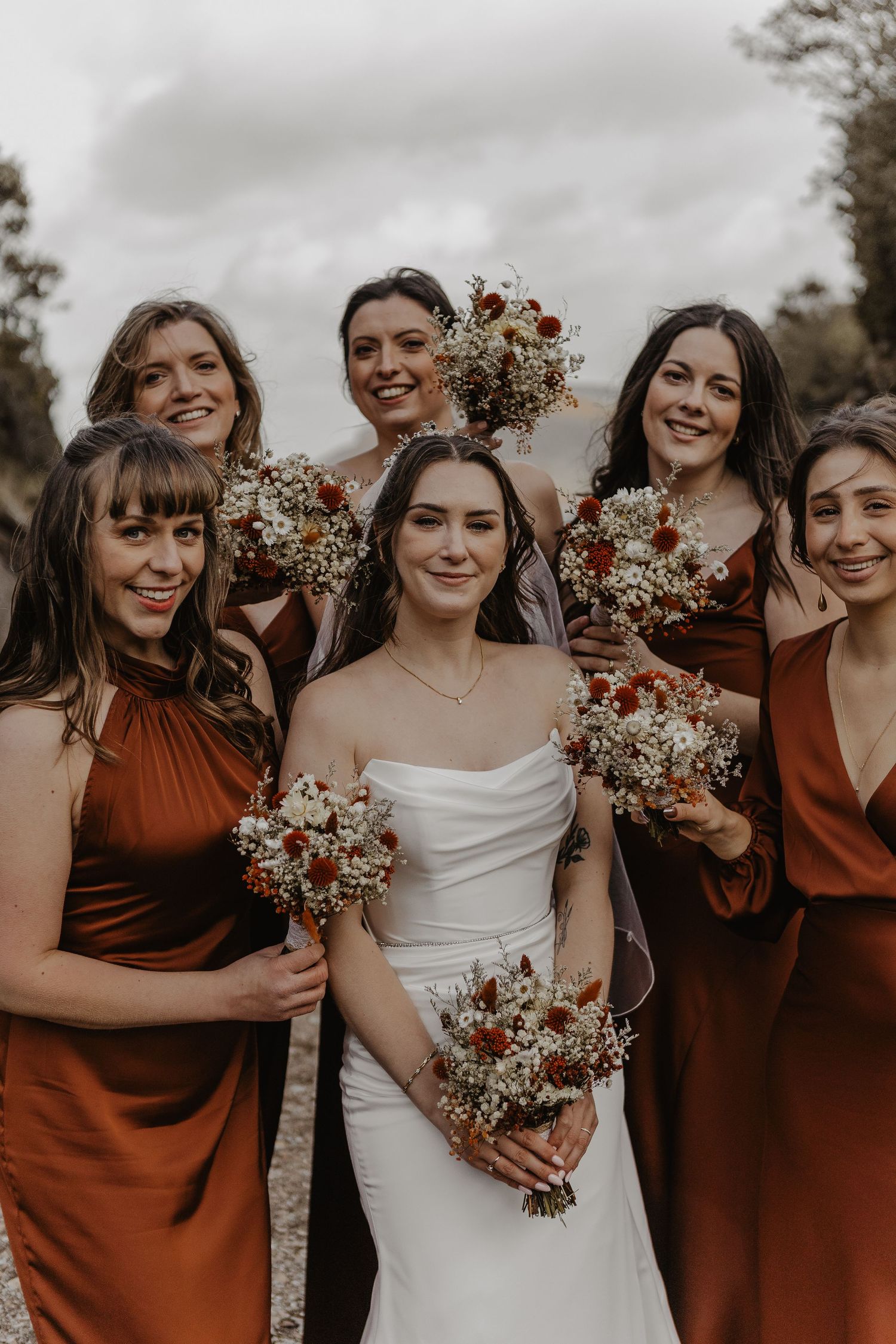 Bridal party in rust-colored dresses holding bouquets against moody sky.