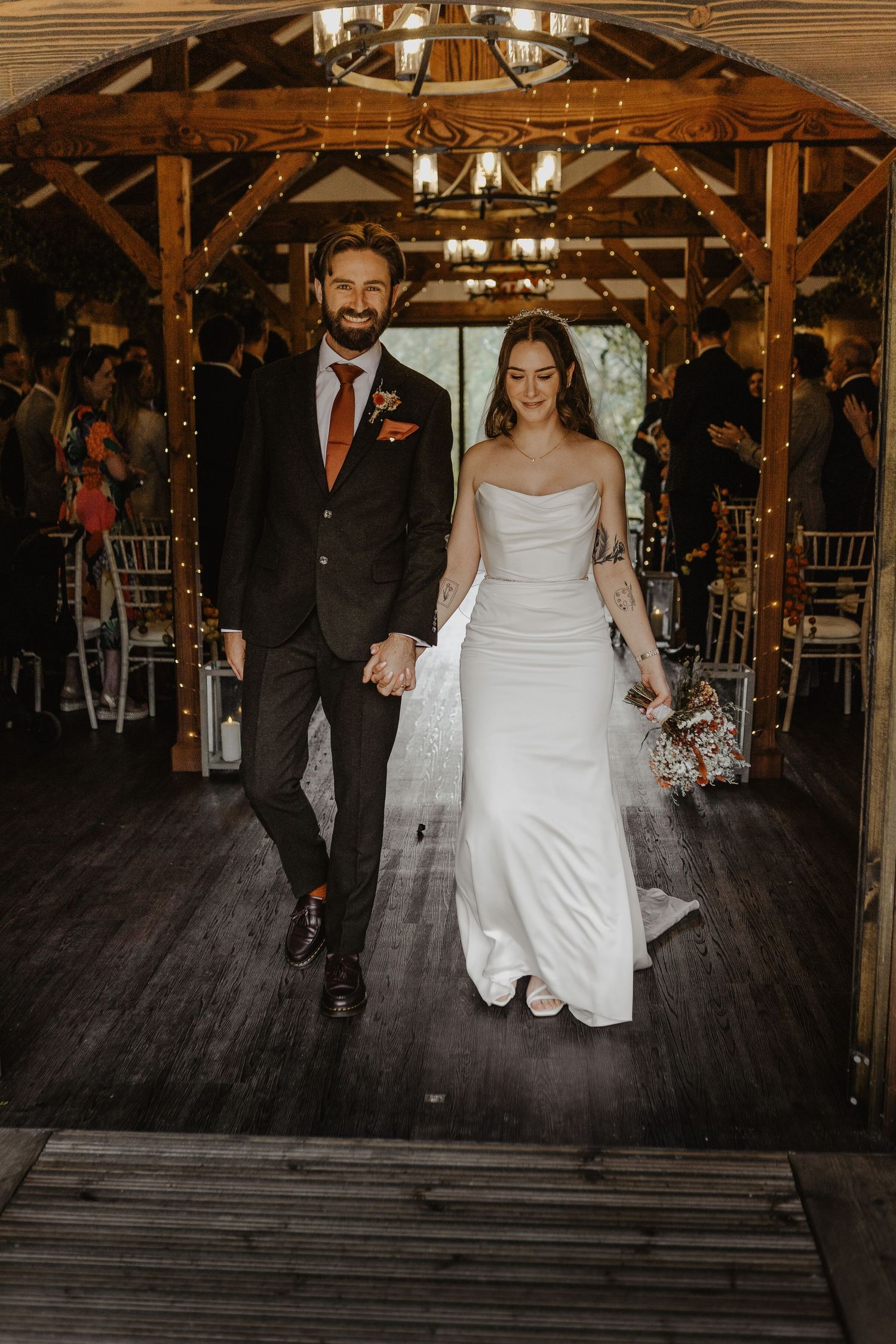 A couple walks across a rustic wooden floor in a barn-style venue decorated with twinkling lights for their wedding.