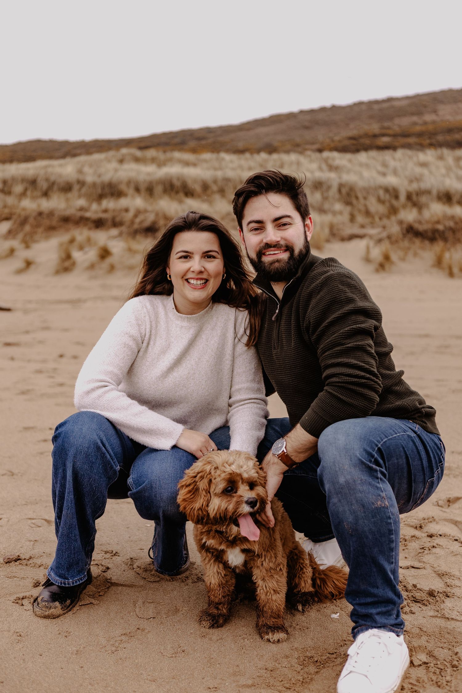 Happy couple posing with their brown dog on a sandy beach with hills in the background.