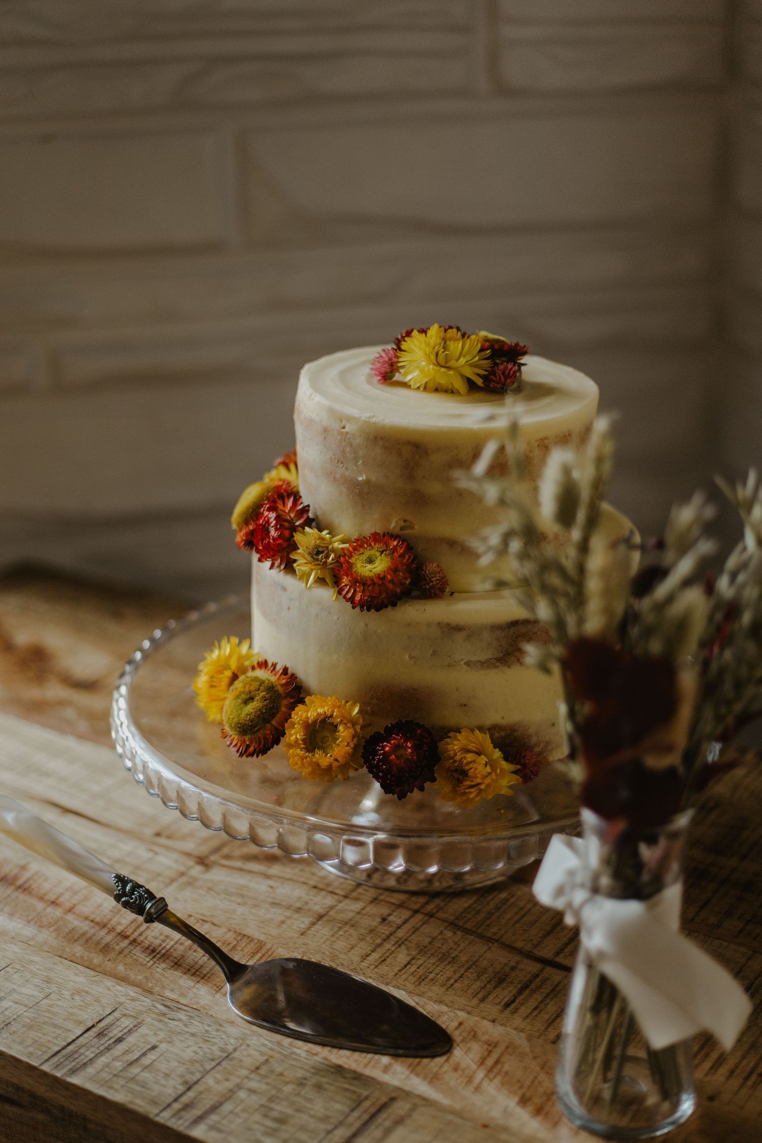 Rustic white frosted cake decorated with yellow and orange dried flowers sits on glass pedestal against wooden backdrop.
