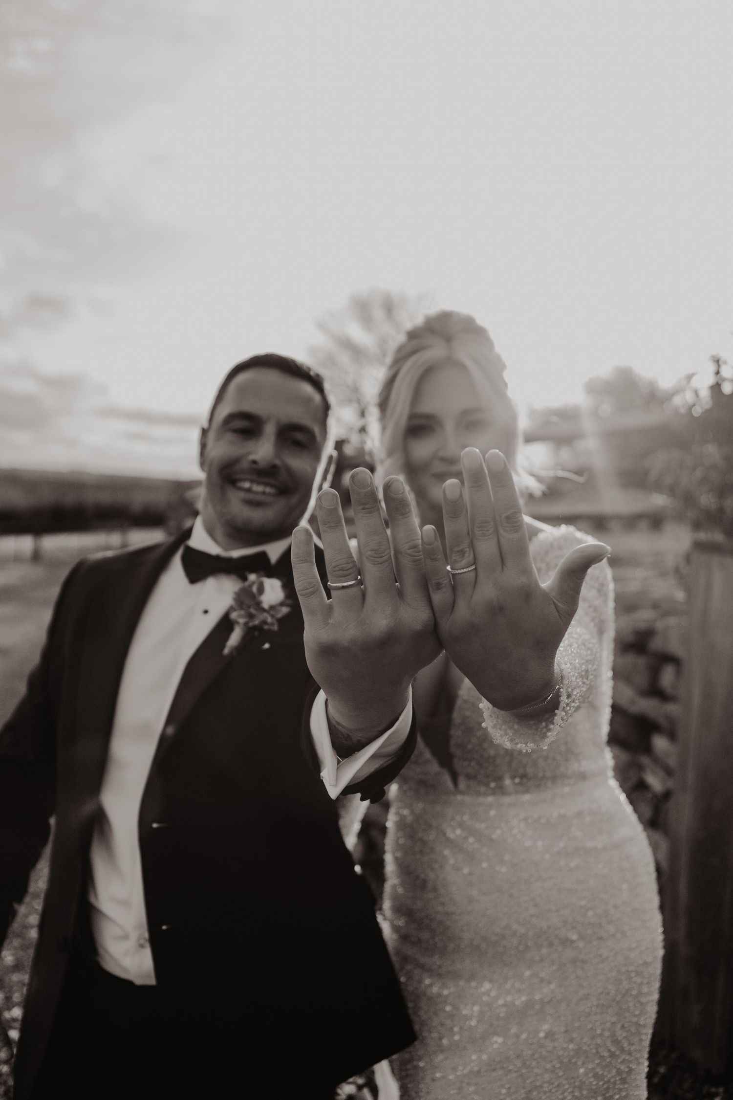 Black and white photo of a newlywed couple showing off their wedding rings with joyful expressions at sunset.