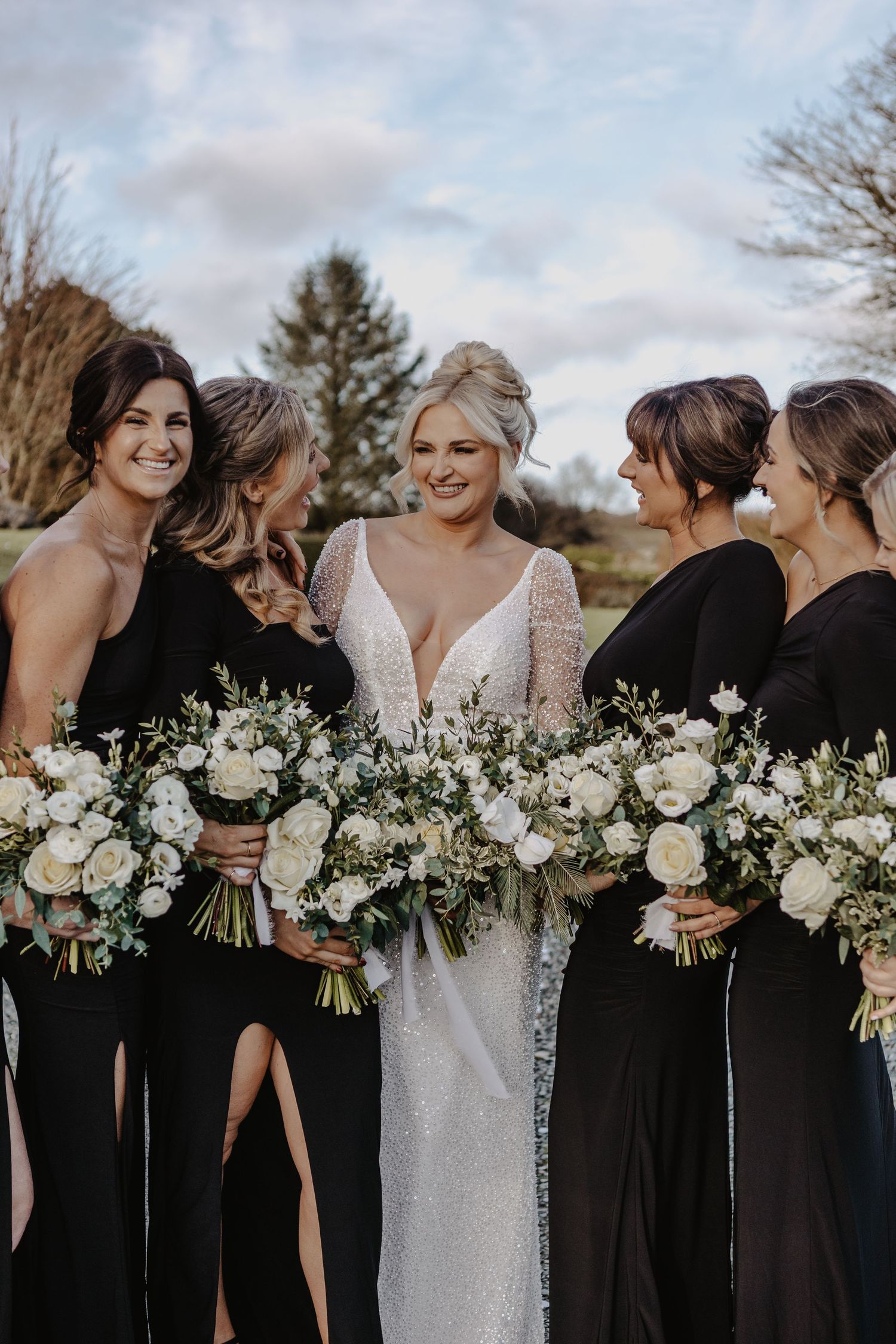 Wedding party in elegant black bridesmaid dresses and white gown hold matching white floral bouquets in outdoor setting.