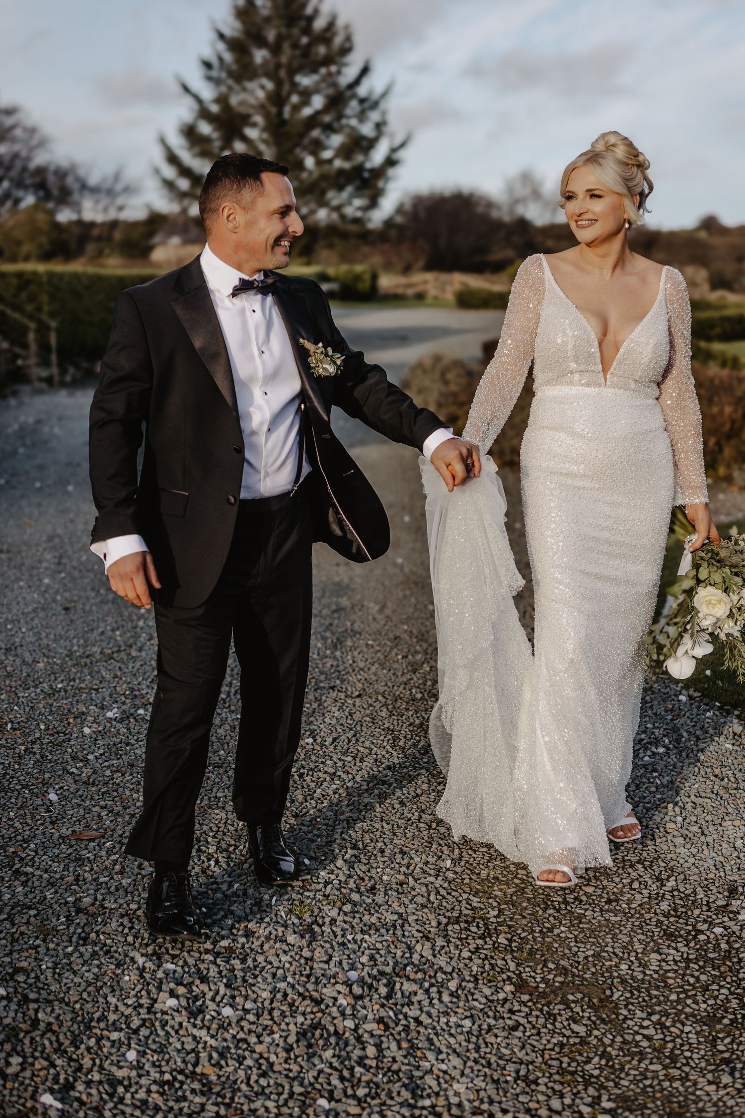 A wedding couple walks on gravel path in formal attire as photographer captures candid moments in sequence.
