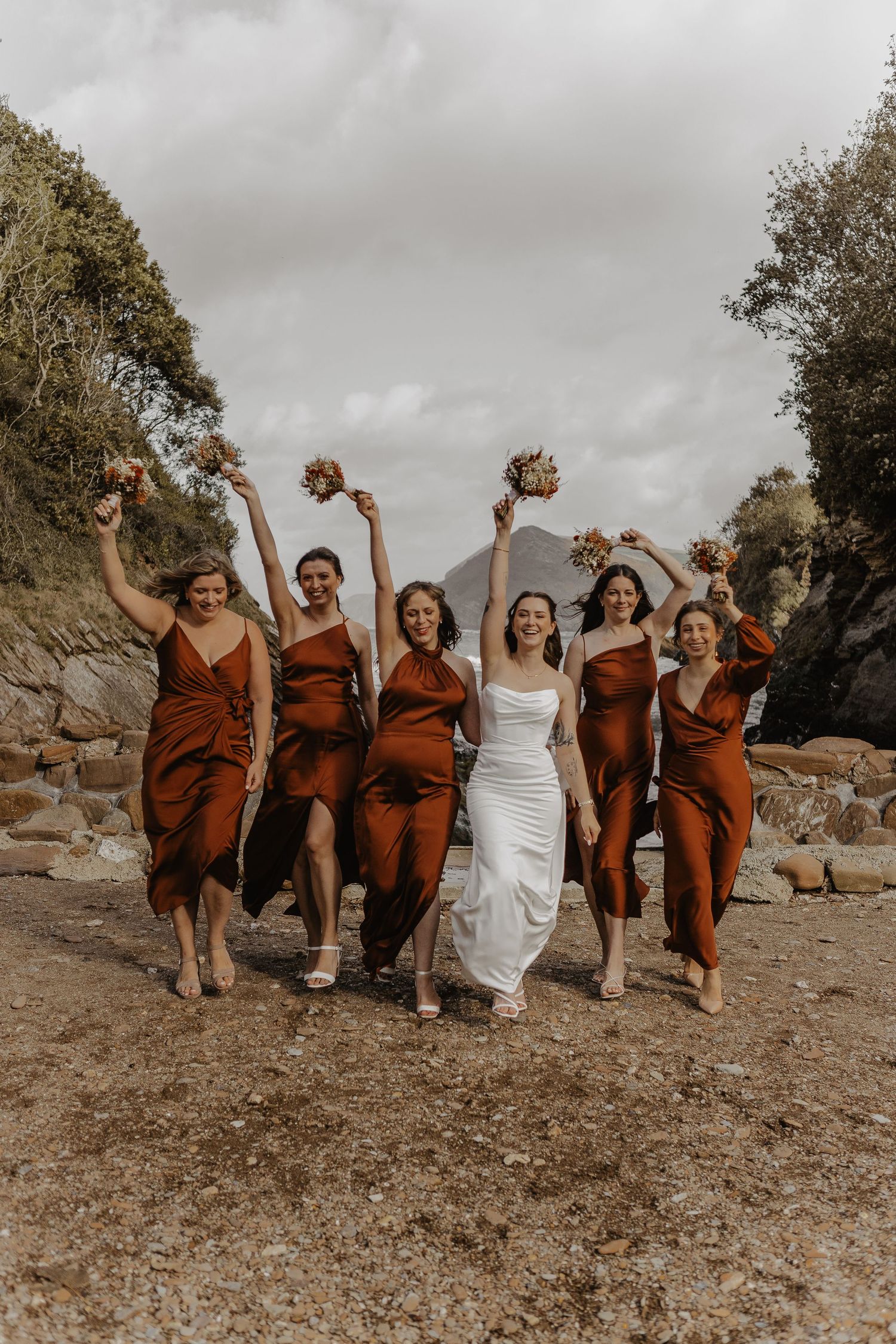 A wedding party in rust-colored dresses poses joyfully on a rocky hillside.