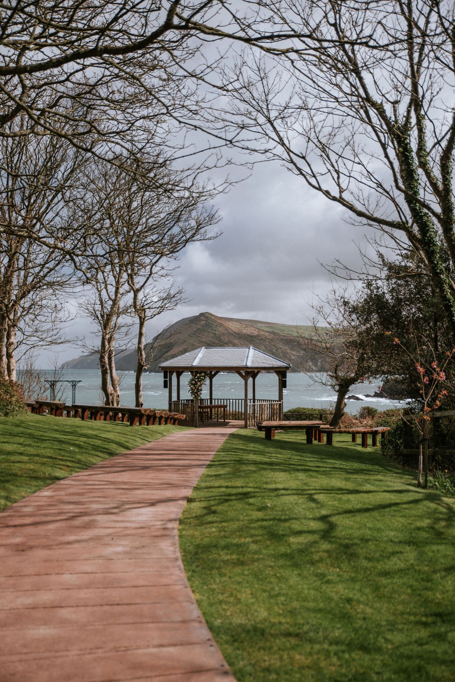A brick pathway leads to a seaside gazebo surrounded by bare trees and green lawns with ocean views.