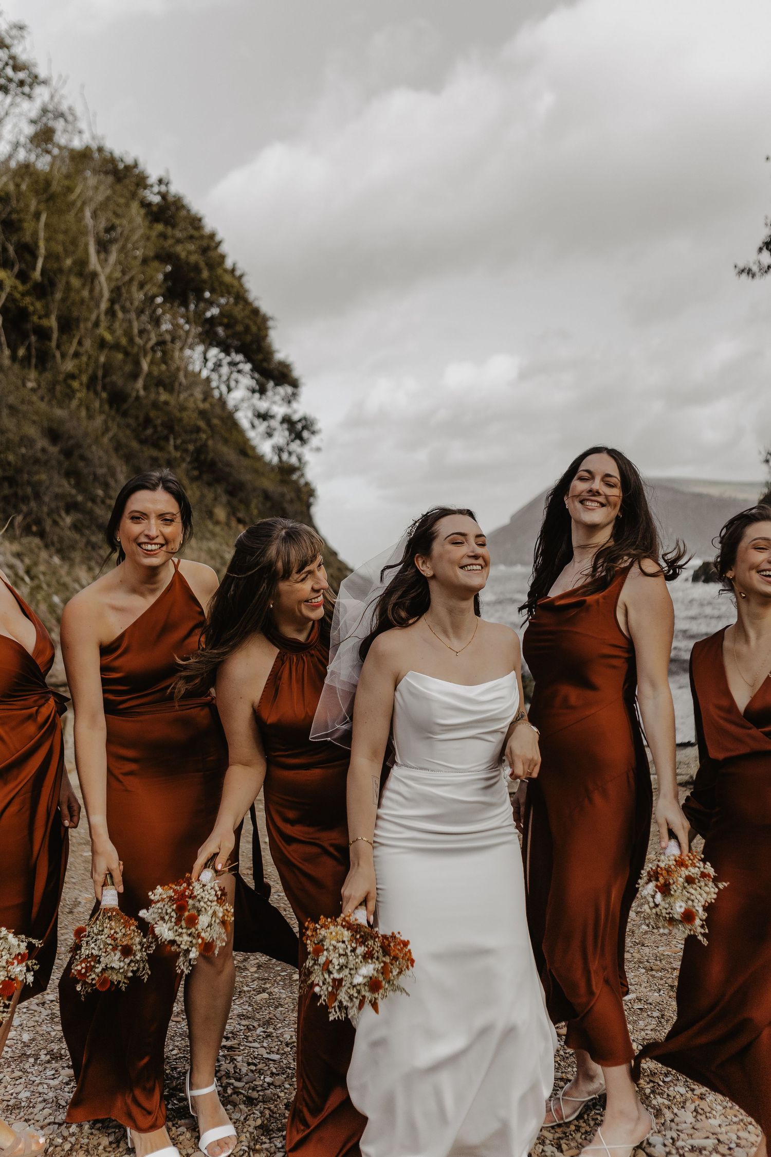 A wedding party in rust-colored bridesmaid dresses and white bridal gown laugh together on a cloudy beach day.